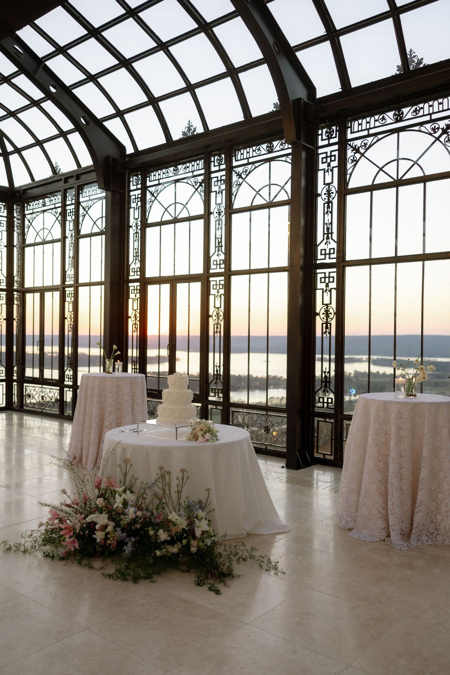 Elegant wedding reception in a glass conservatory with a tiered cake, floral arrangements, and stunning sunset water views.