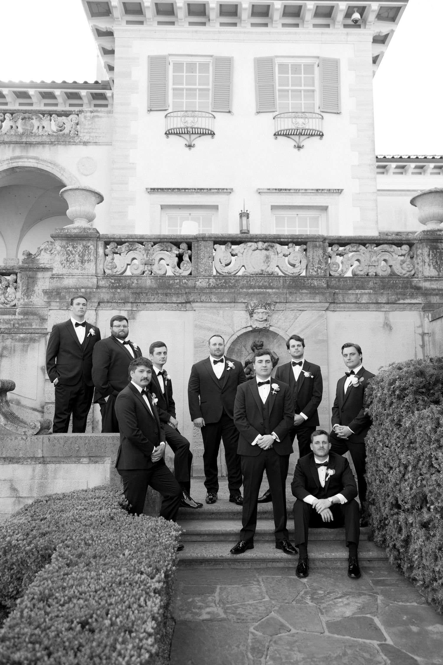 Groom and groomsmen in black tuxedos pose on ornate stone steps of a grand estate in black and white.