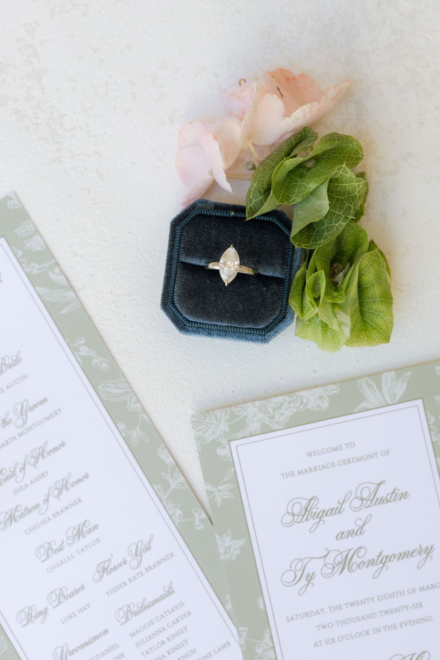 Marquise diamond engagement ring in velvet box beside sage green wedding program and pink roses.