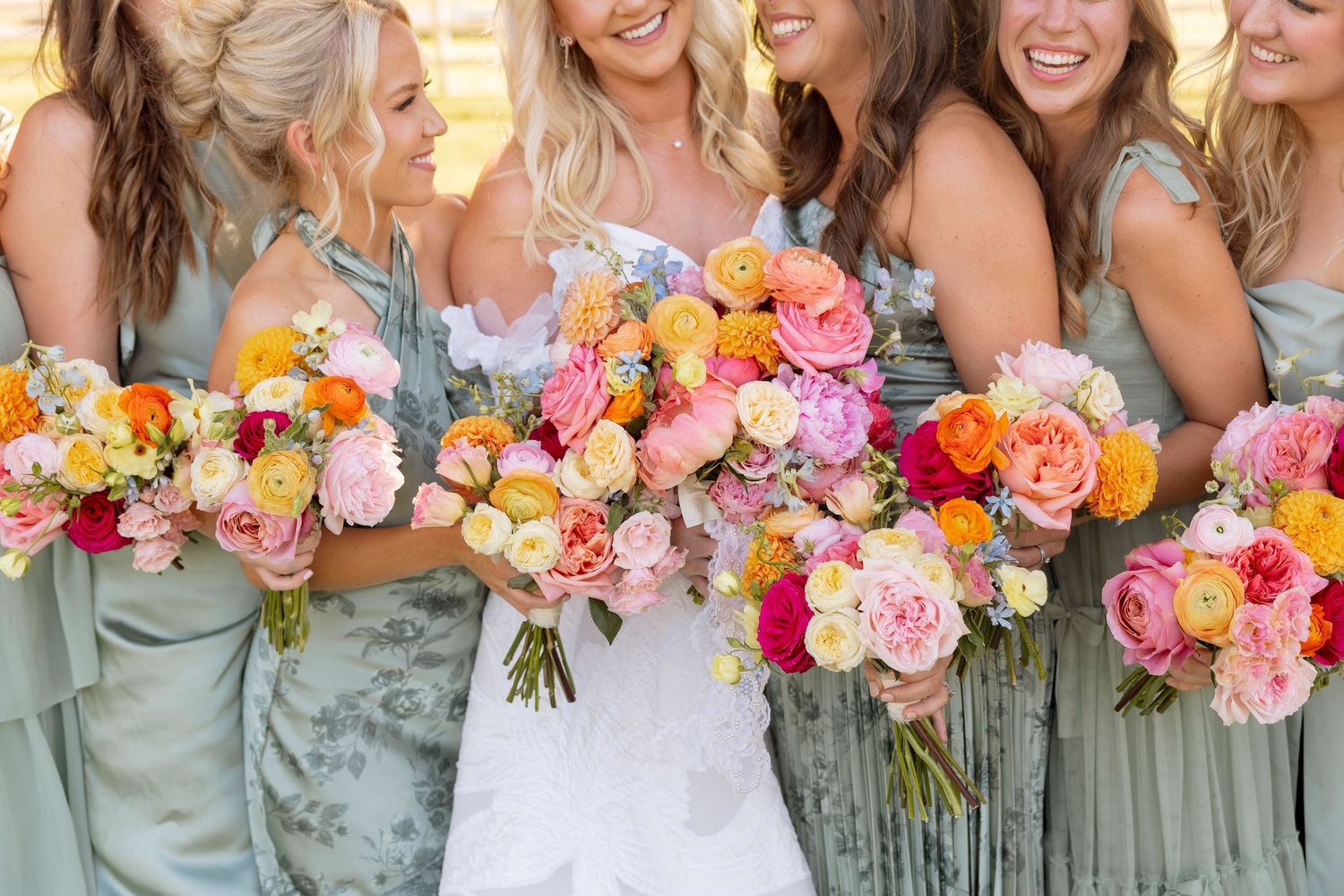 Bridesmaids in sage green dresses hold vibrant pink and orange bouquets while smiling.
