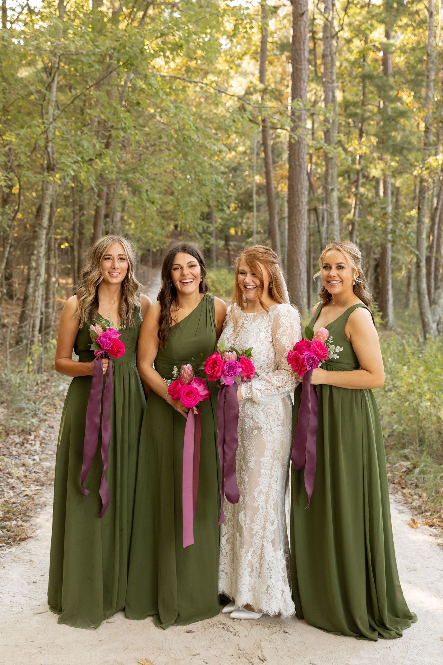 Wedding portrait of four women in green dresses and white lace gown holding pink bouquets outdoors.