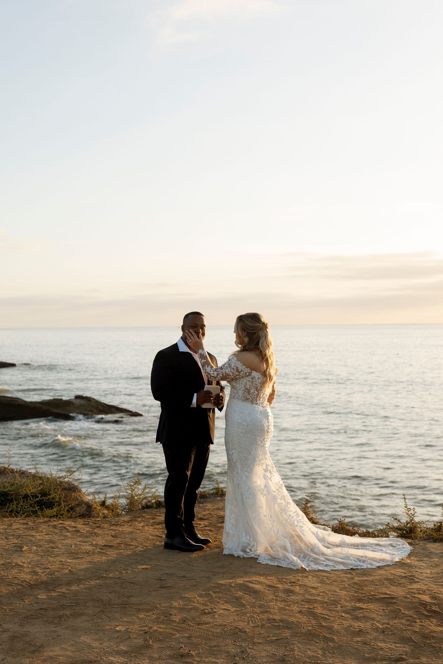 A couple in wedding attire shares an intimate moment on a rocky coastal cliff at sunset by the ocean.