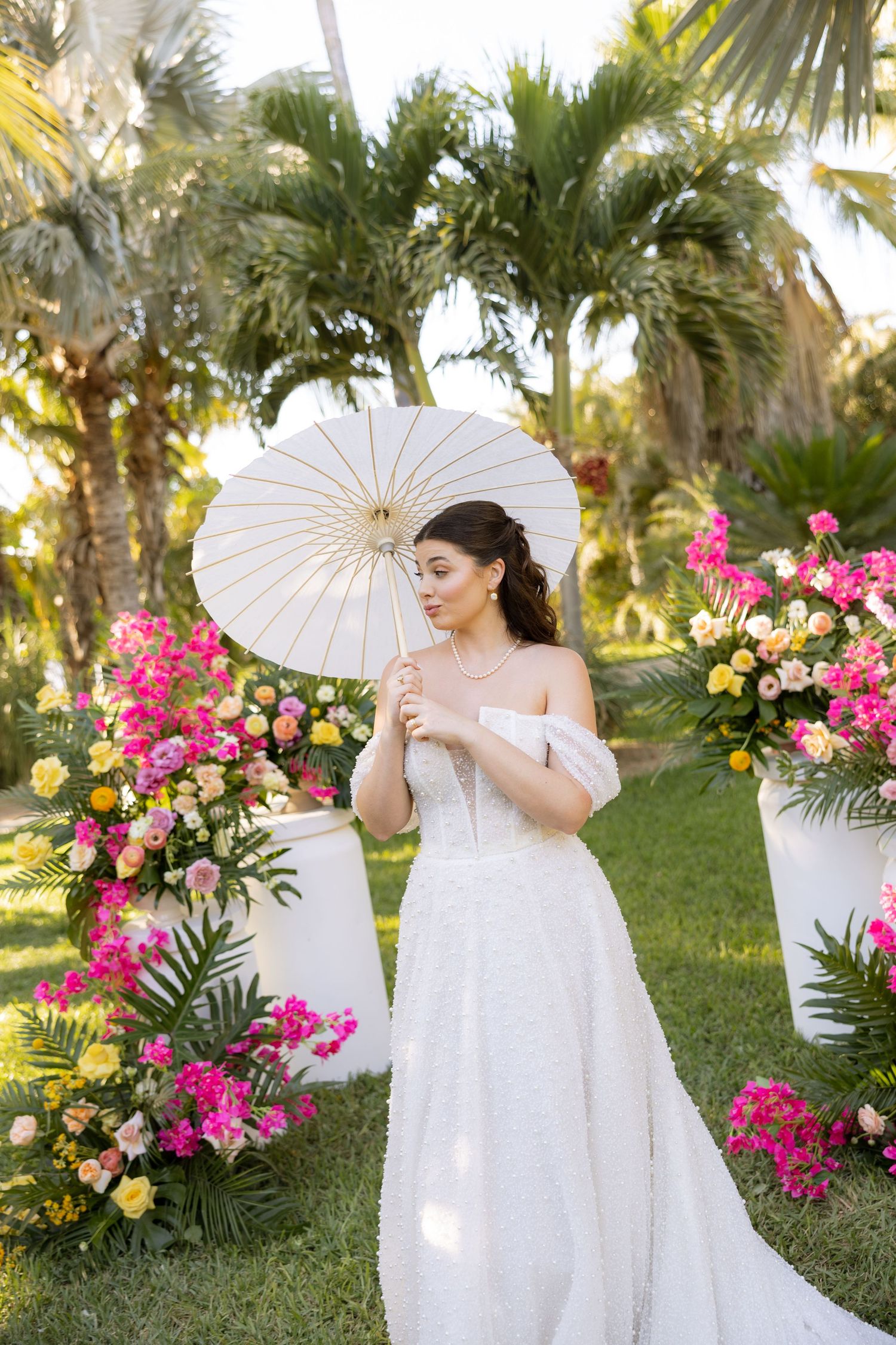 A bride in a lace gown holds a white parasol surrounded by vibrant tropical floral arrangements.