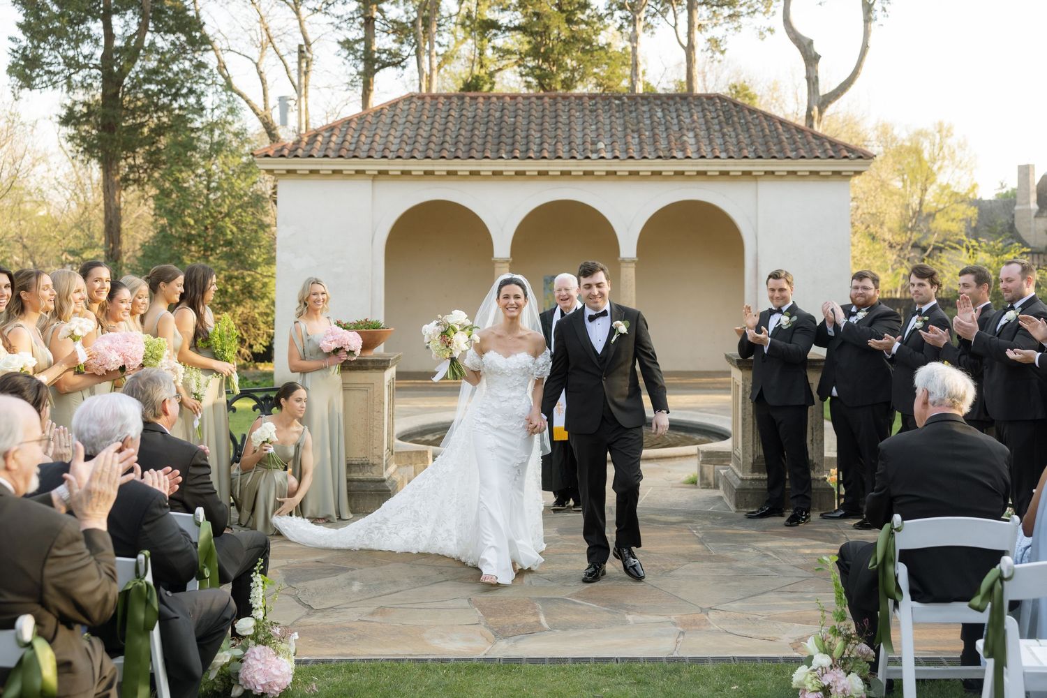 Bride and groom walk down outdoor aisle at elegant garden wedding ceremony with wedding party.