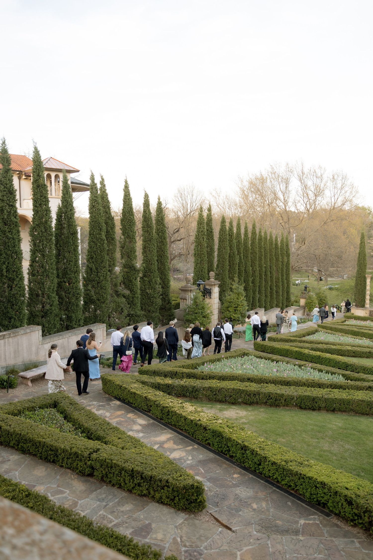 Wedding guests stroll through manicured Italian garden with tall cypress trees at elegant venue.