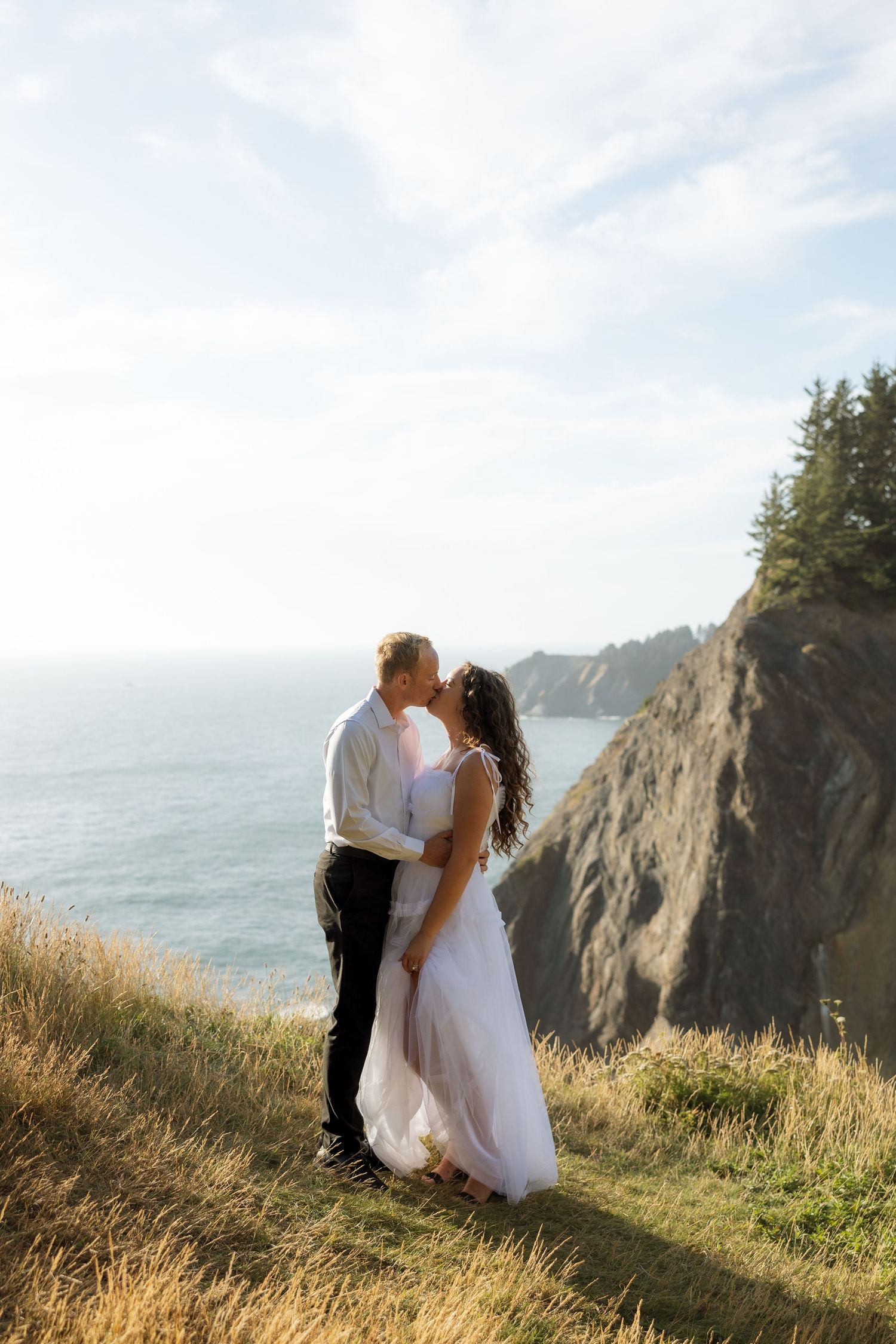 Newlyweds embrace on a scenic coastal clifftop with ocean and mountains in the background.