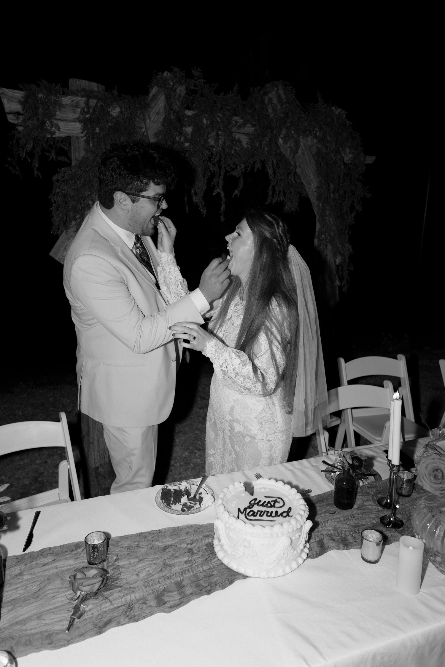 A black and white photo of newlyweds sharing cake at their reception table.