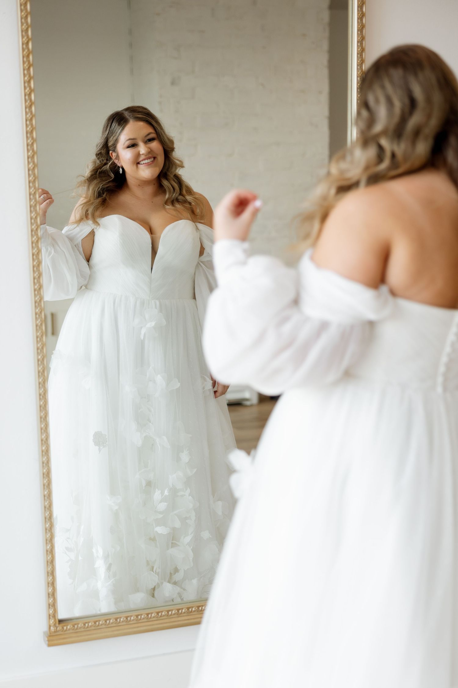 A radiant bride in an off-shoulder wedding dress admires herself in a gold-framed mirror.