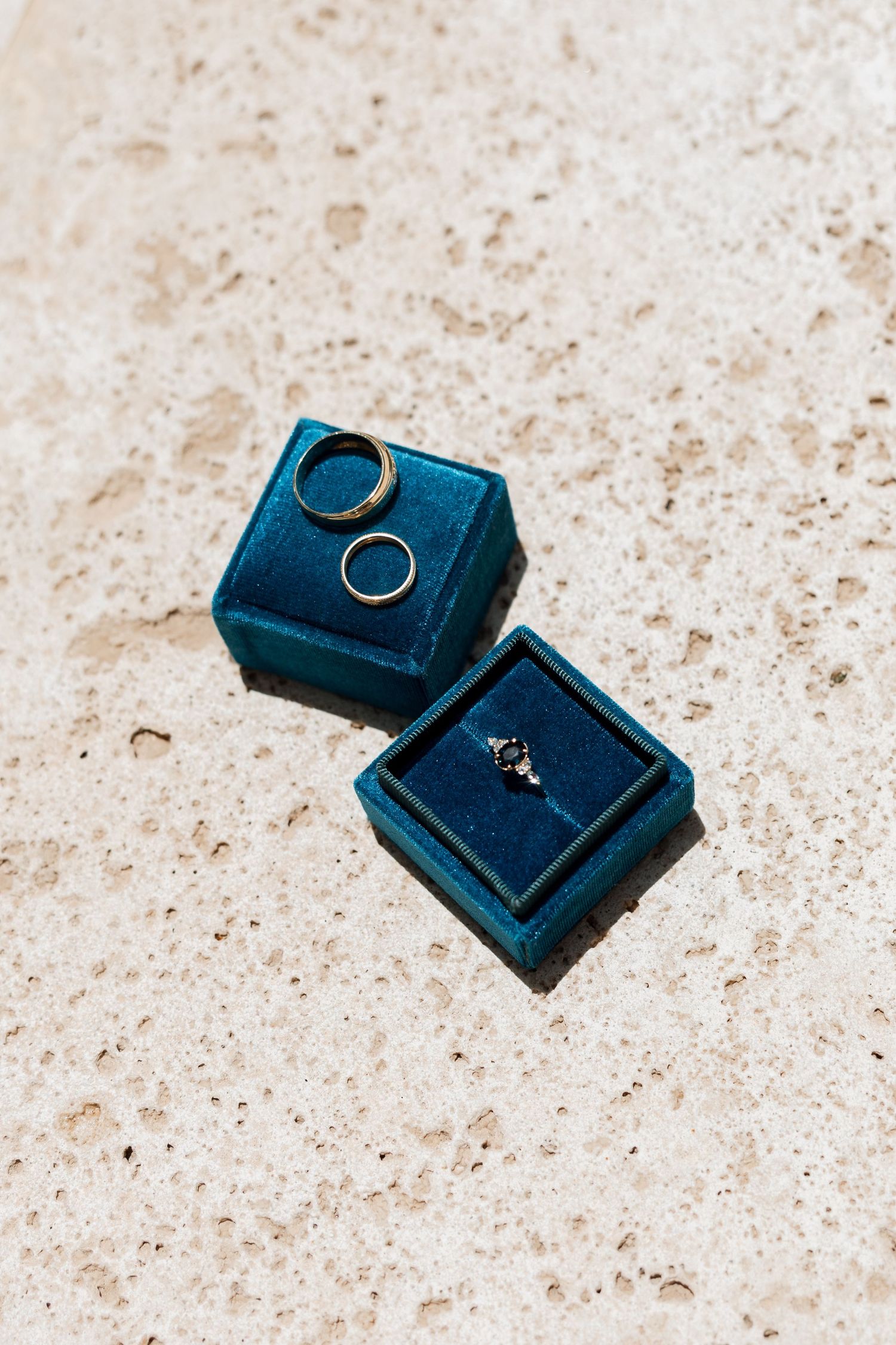 Wedding rings displayed in a dark teal velvet ring box on a light marble surface.