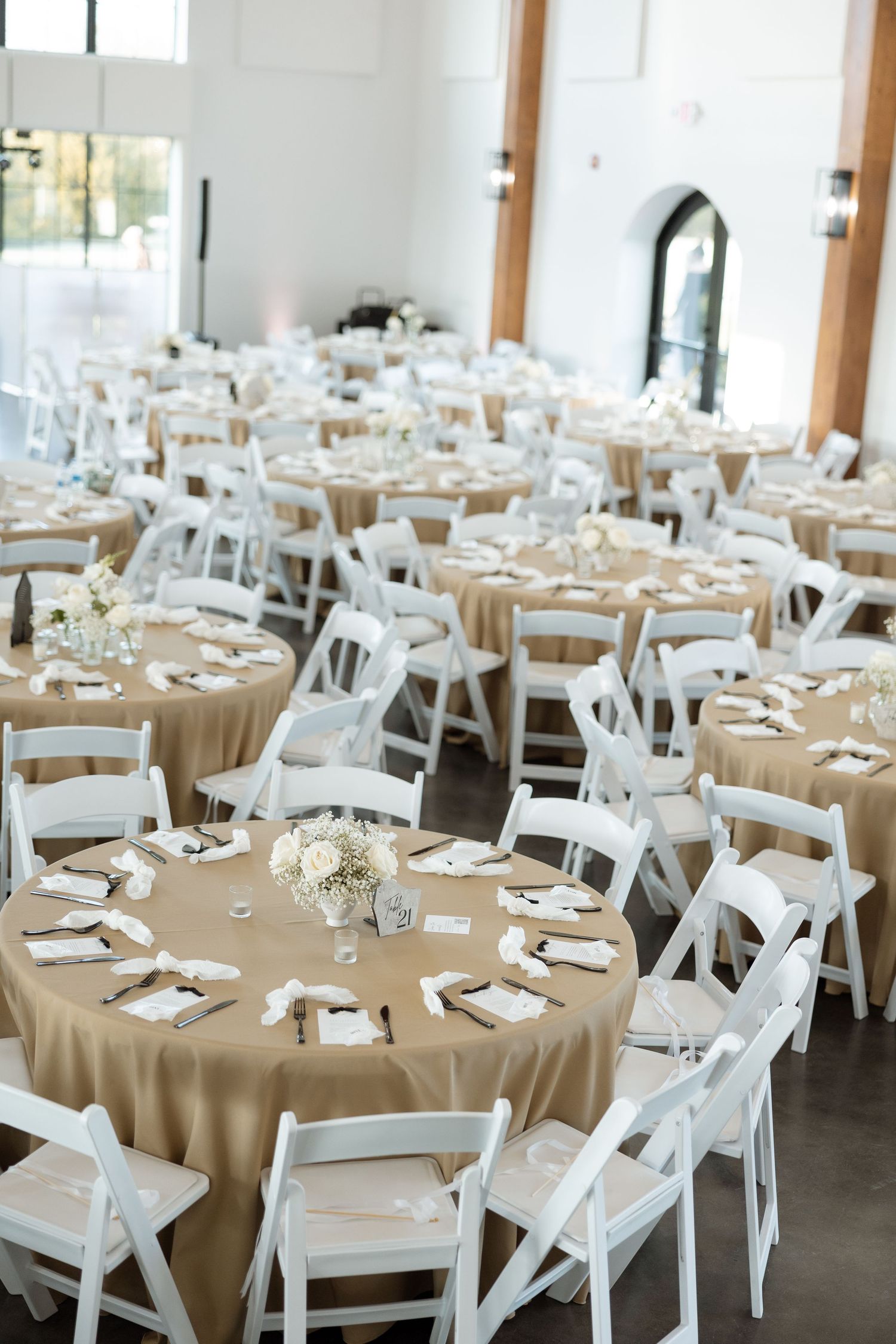 Wedding reception venue with round tables set with tan tablecloths and white folding chairs.