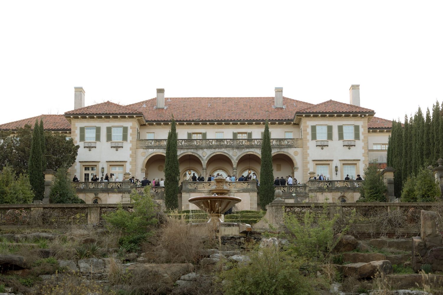 Grand Italian-style villa with arched colonnade, terracotta roof, fountain, and cypress trees at wedding venue.