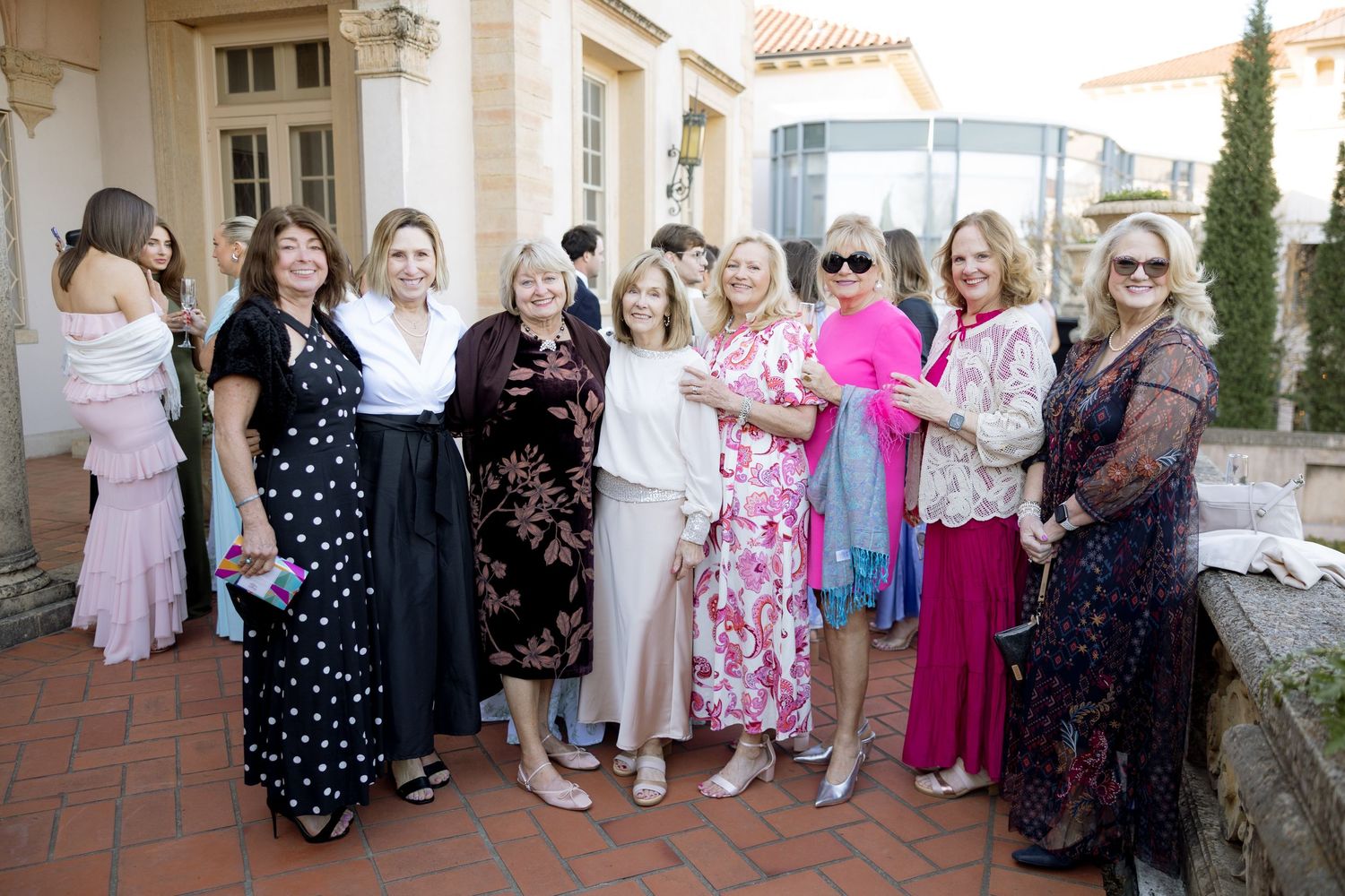 Group of elegantly dressed women smiling together on terrace at outdoor wedding reception.