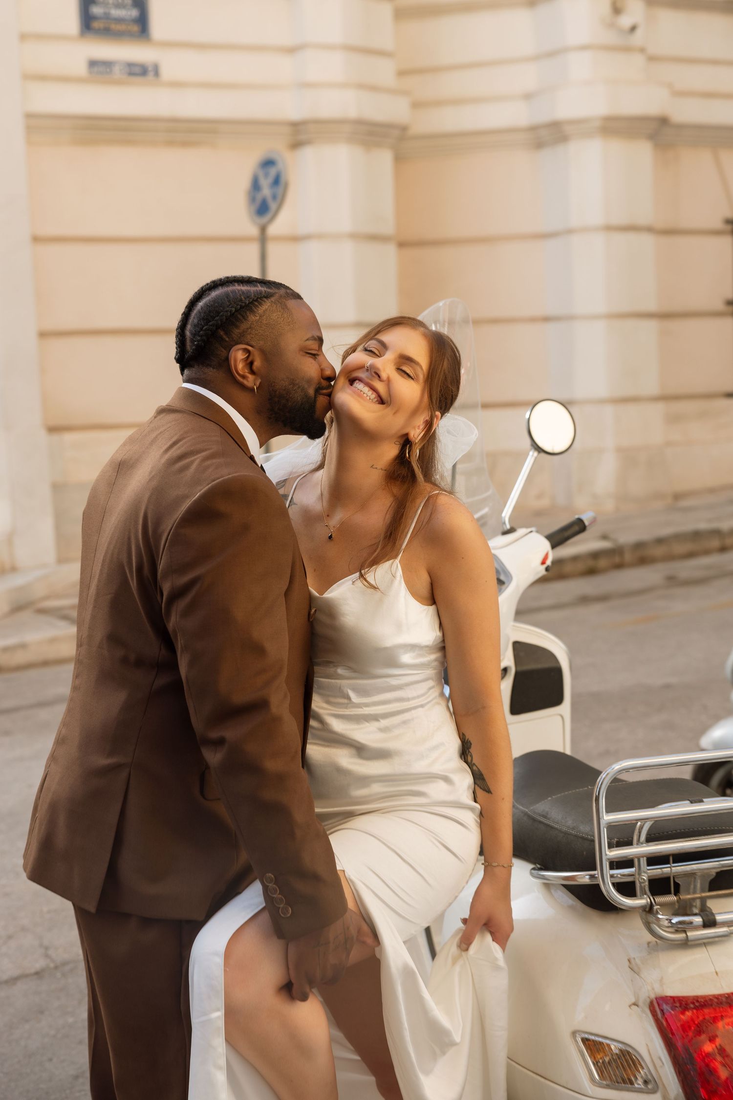 Intimate moment shared between newlyweds next to a white vintage scooter on city street.