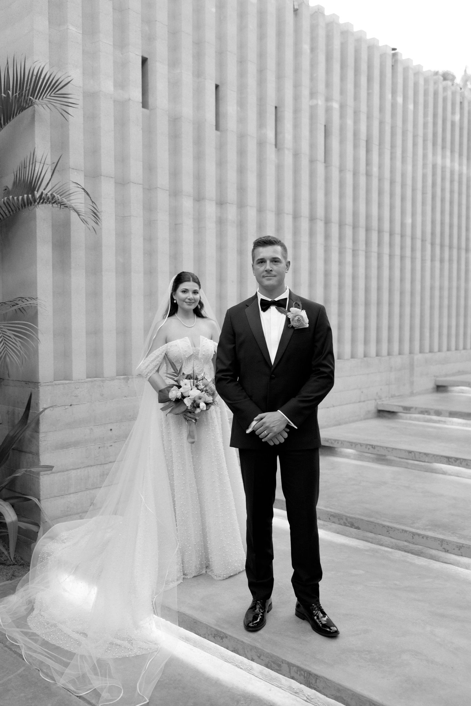 Bride in off-shoulder gown with long veil and groom in tuxedo pose by stone columns in black and white.