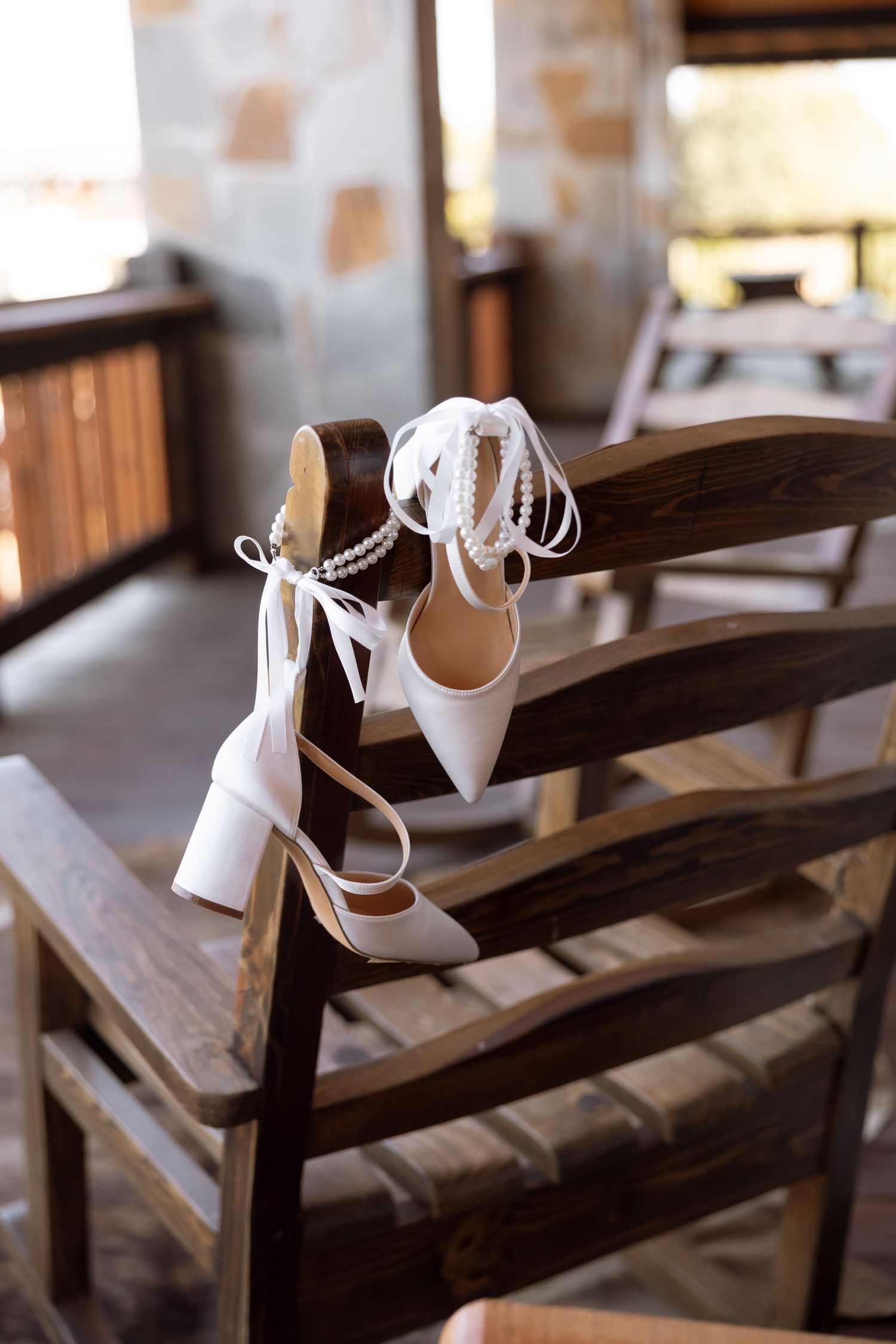 Elegant white wedding shoes with ankle straps displayed on a wooden chair.
