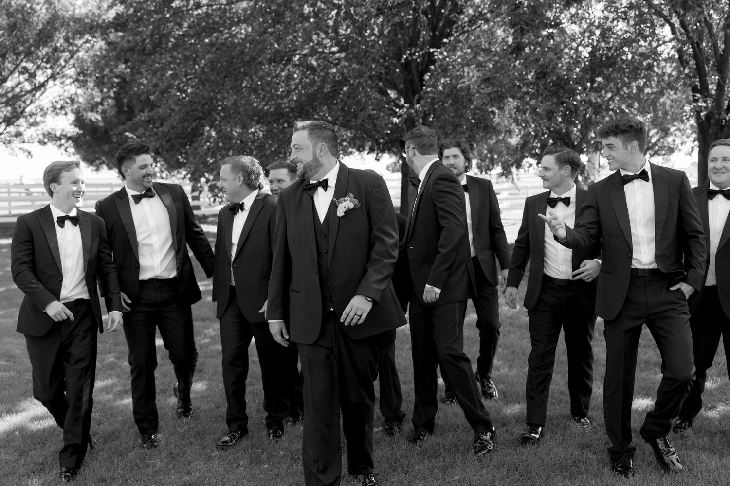 Groomsmen in black tuxedos walking in formation outdoors under tree canopy.