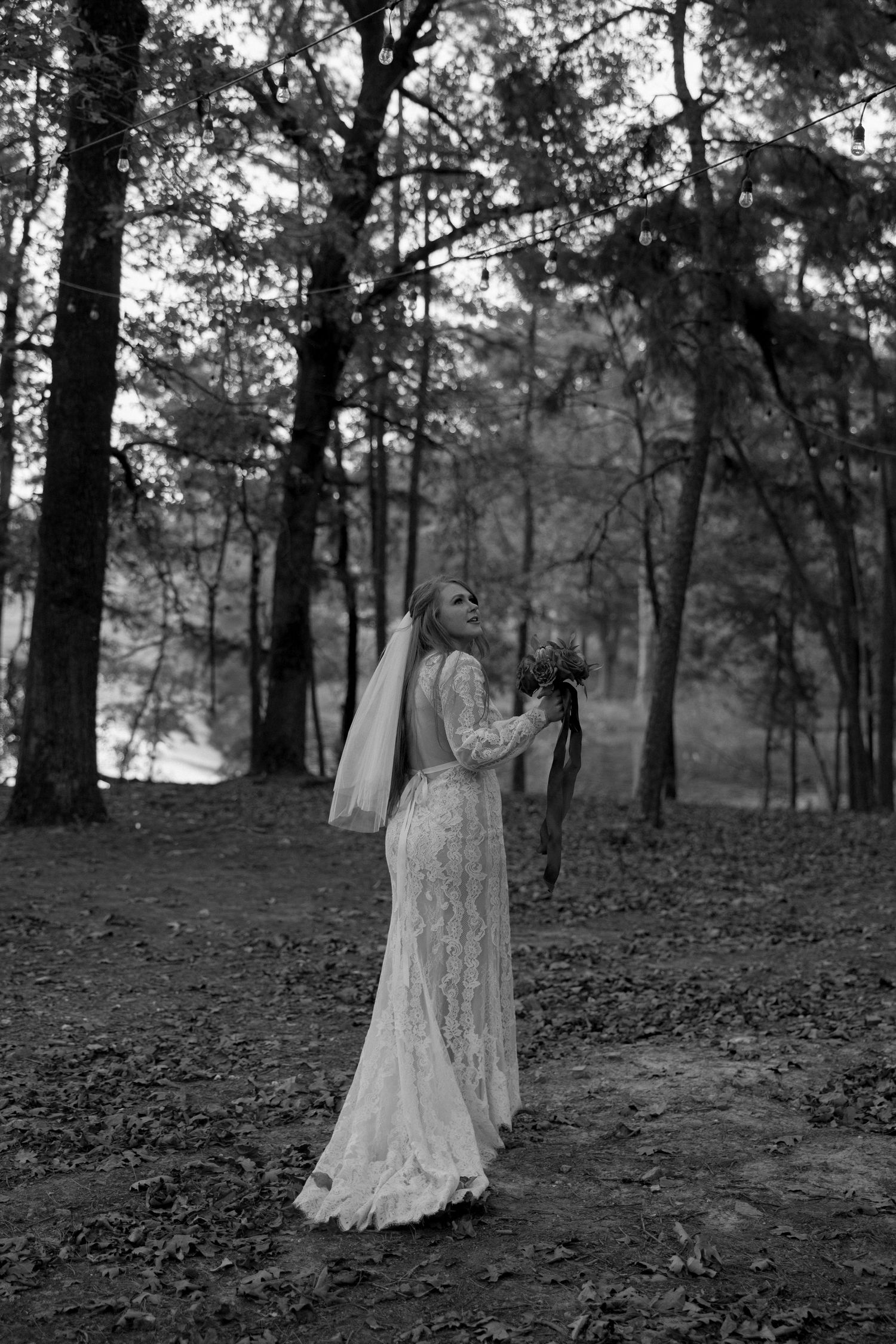 A dramatic black and white portrait of a bride in a lace gown among forest trees.