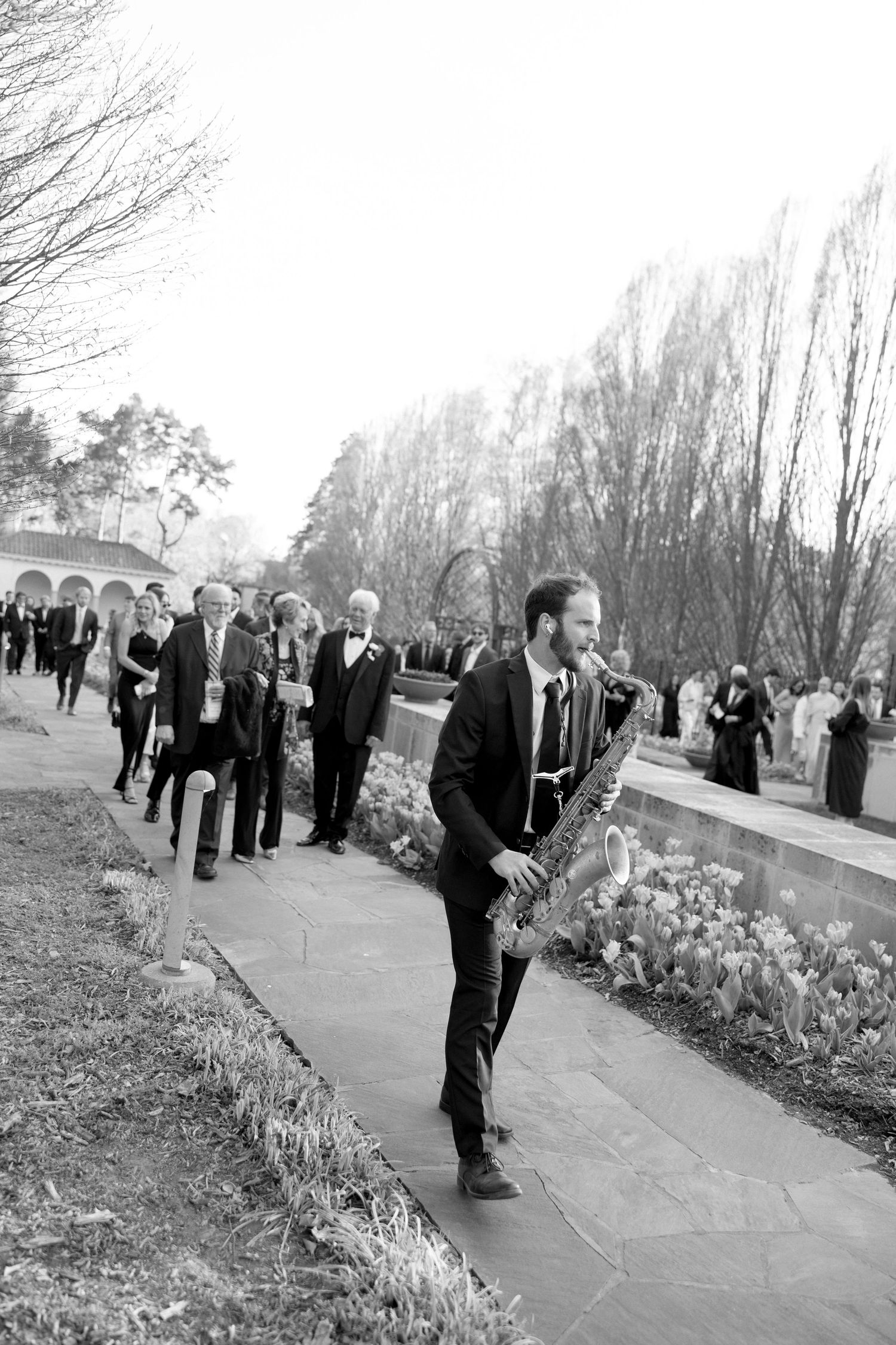 Saxophonist leads wedding guests along garden pathway during outdoor celebration in black and white.