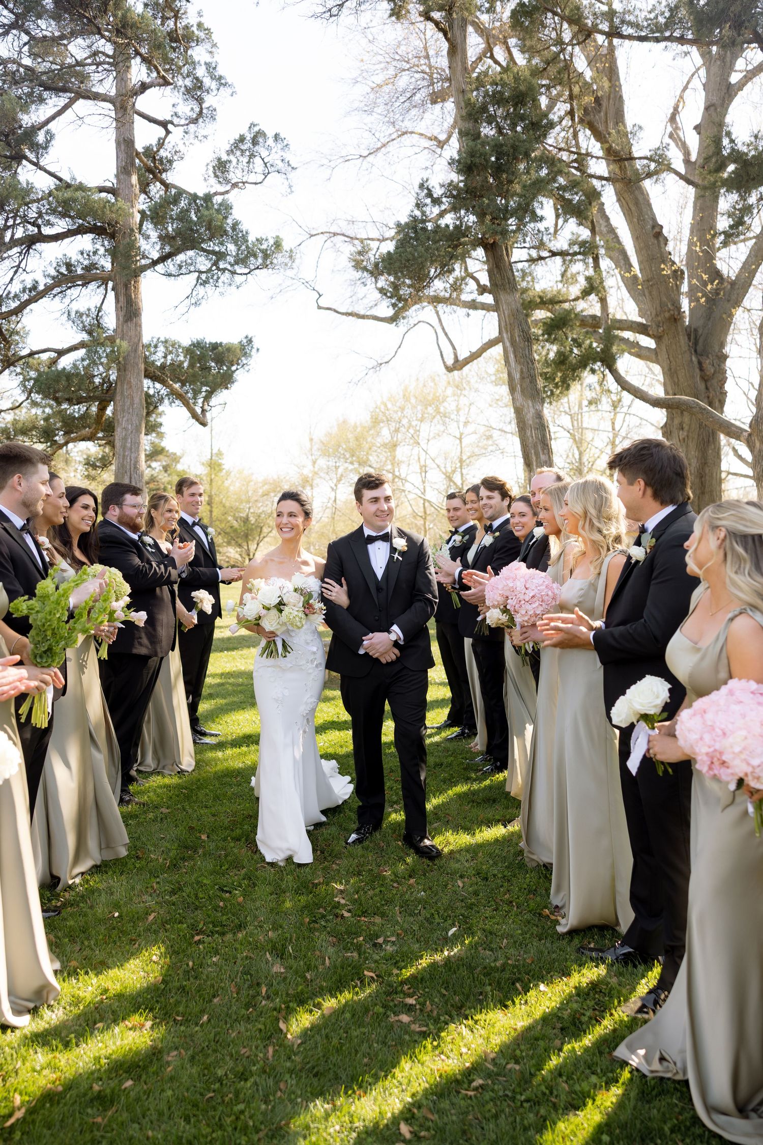 Bride and groom walk down outdoor aisle lined with wedding party in champagne dresses and black tuxedos.