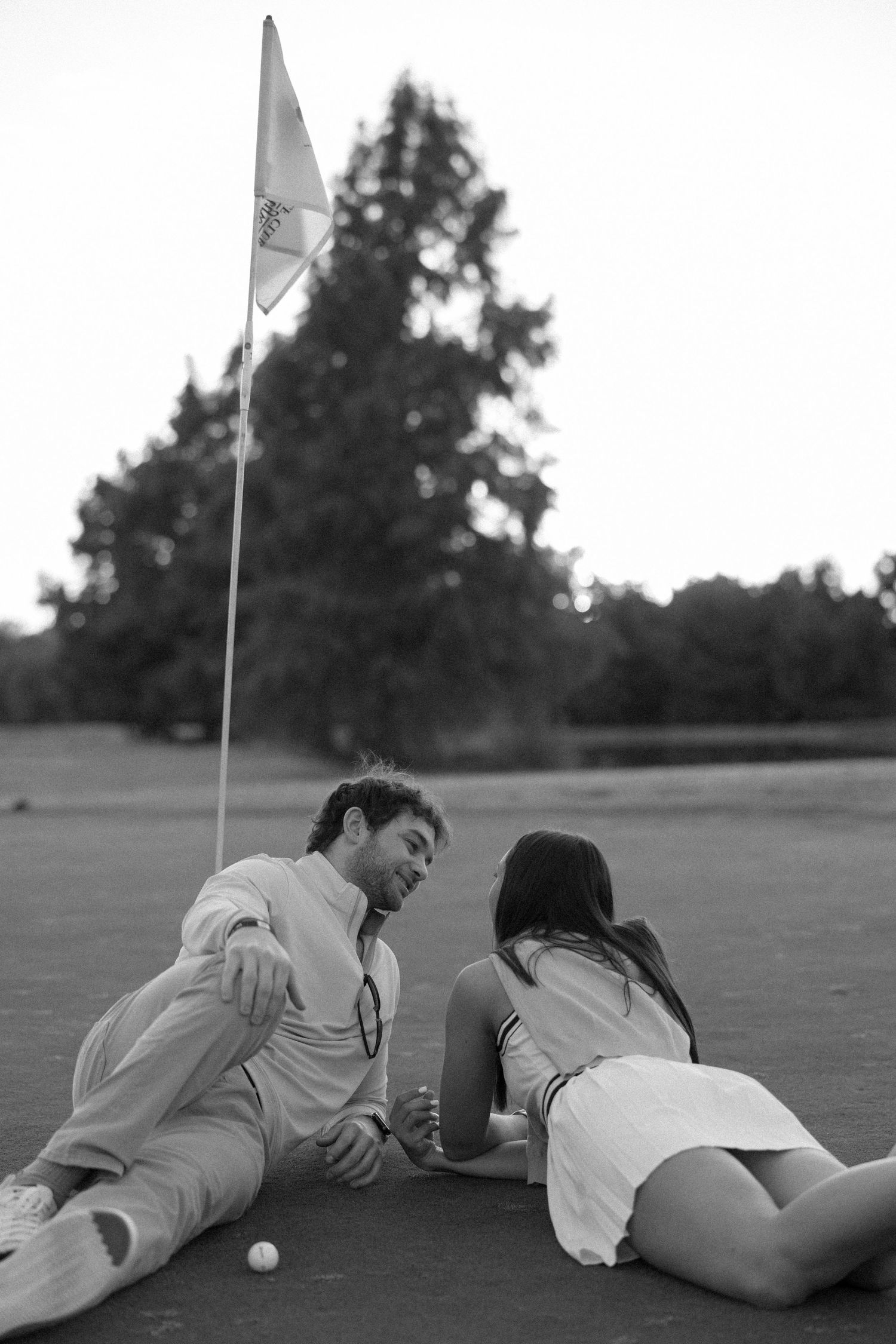 Black and white photo of a couple lying on a golf green next to a flag pole.