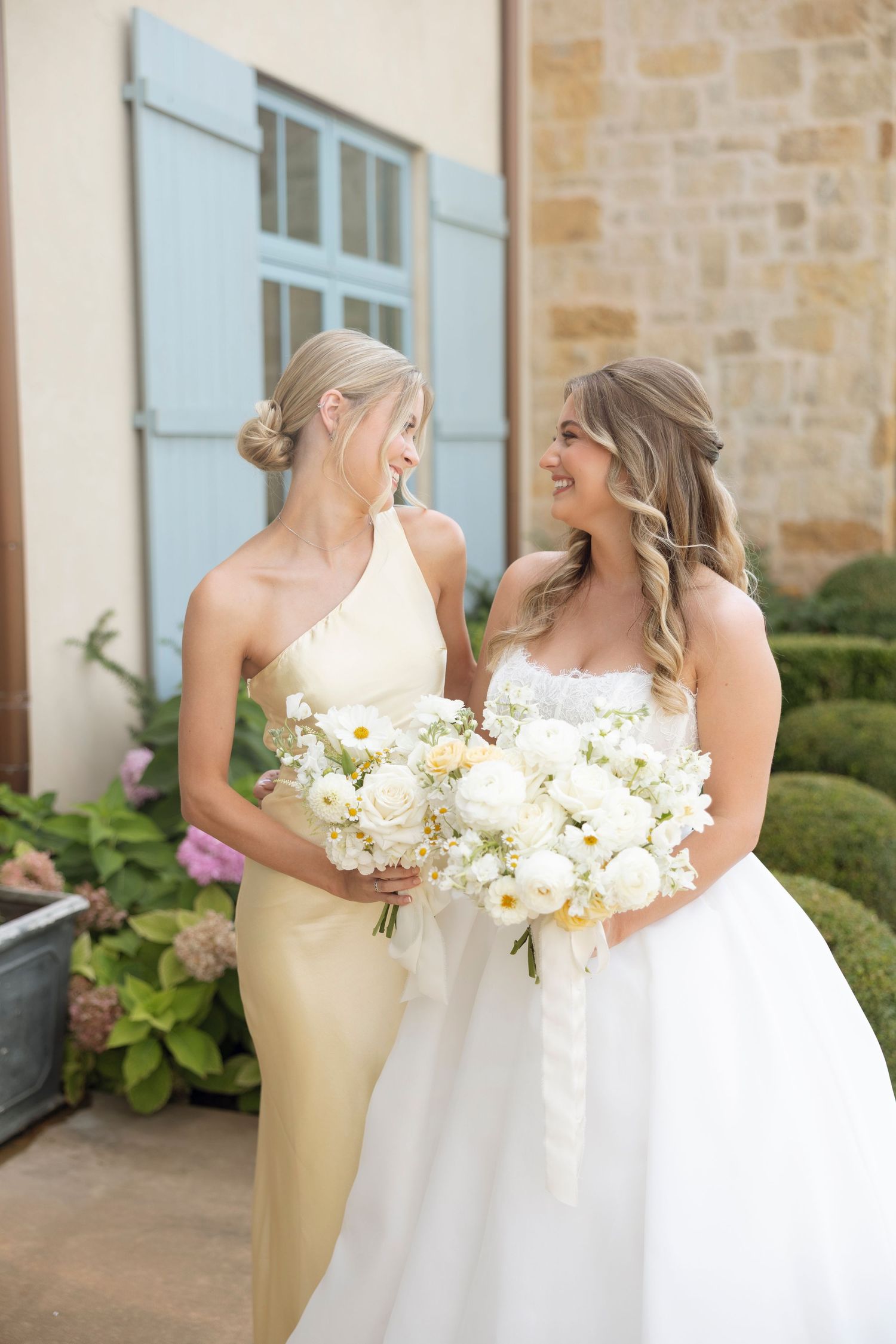 Two women share a candid moment while holding white bouquets against a stone wall with blue window shutters.