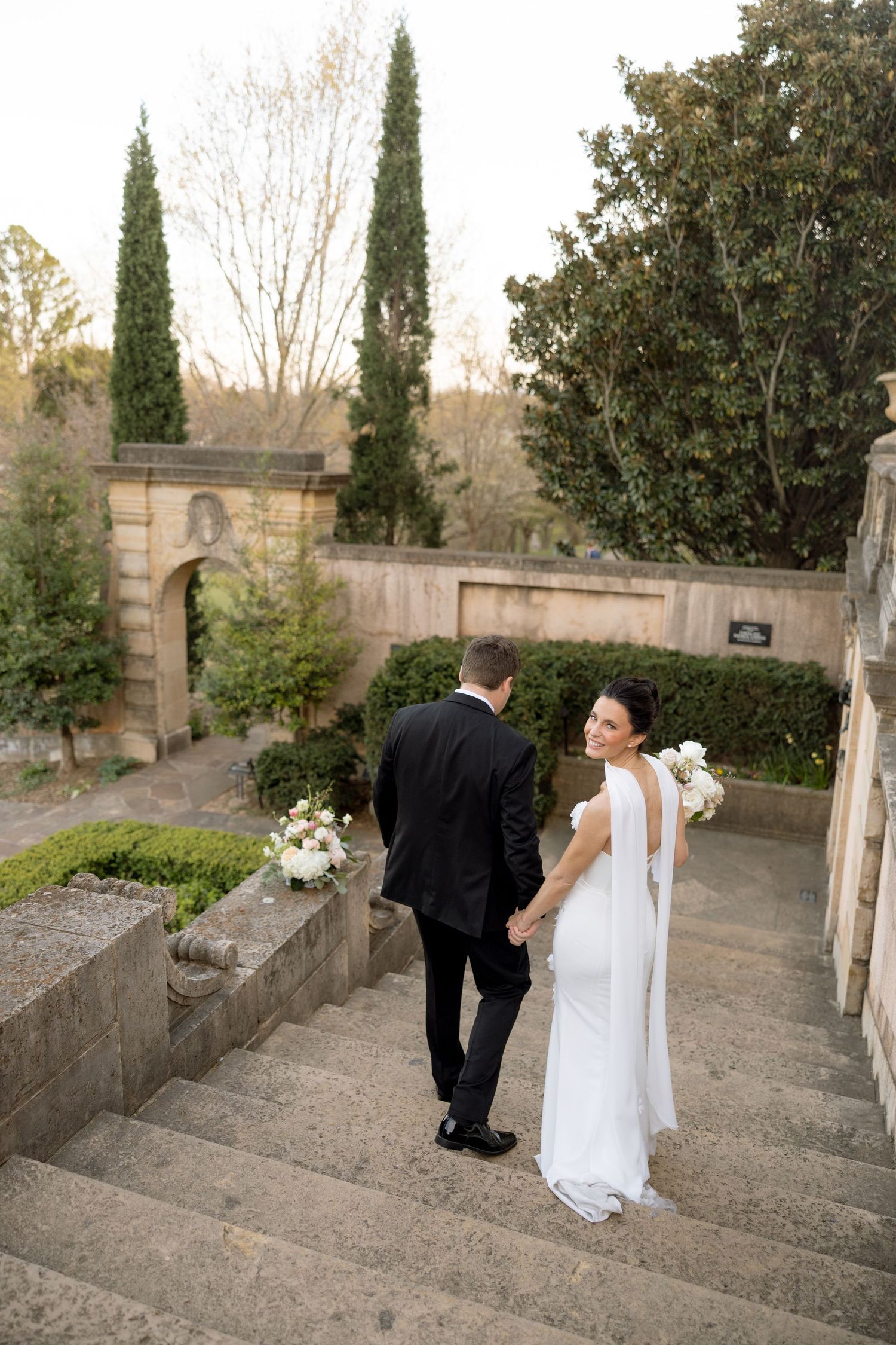 Bride and groom walk hand-in-hand up stone steps in a formal garden with cypress trees and stone archway.