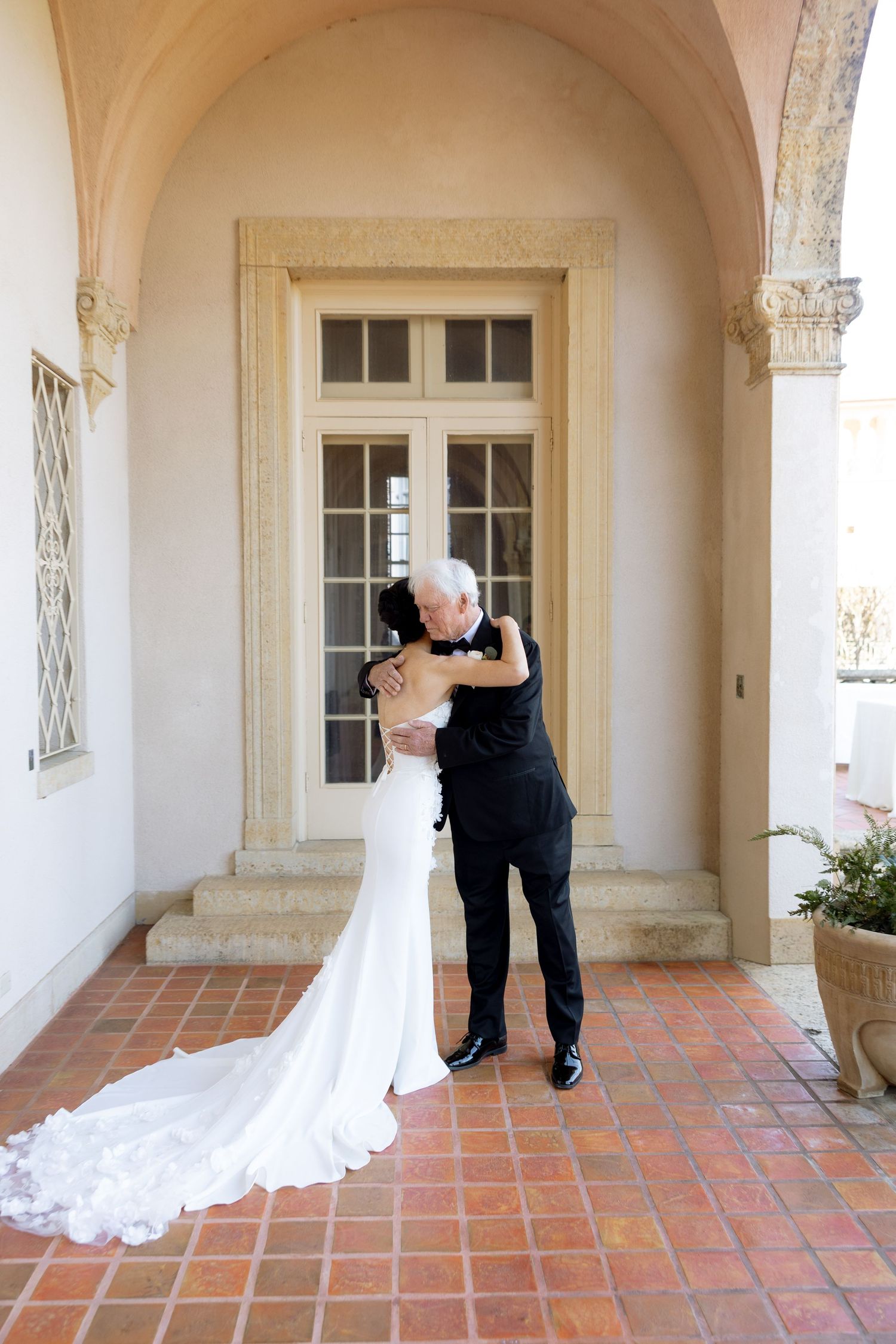 Bride in fitted white gown sharing emotional hug with older gentleman in tuxedo at elegant venue.