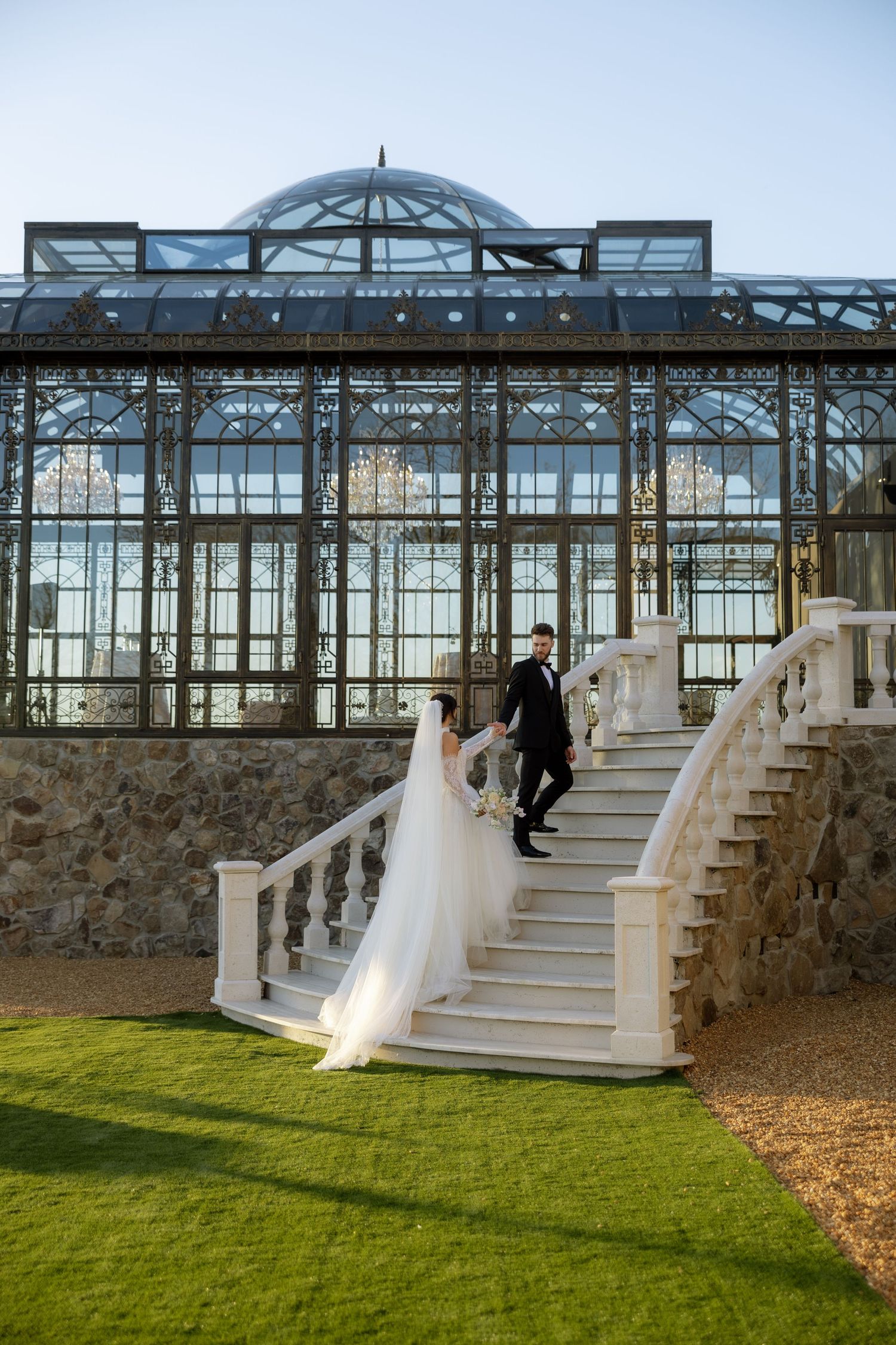 Bride and groom hold hands on elegant white stone staircase before a grand glass greenhouse wedding venue.