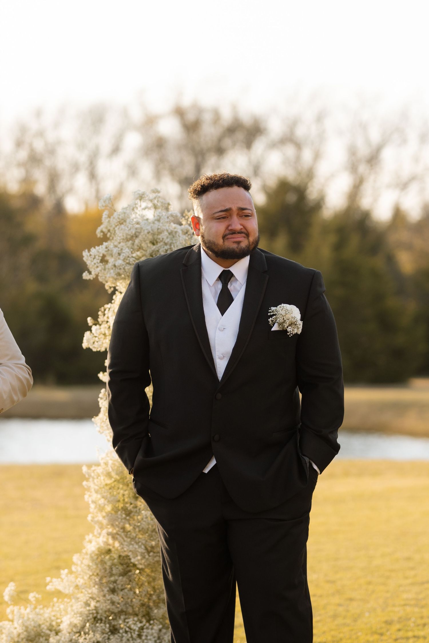 Groom in classic black tuxedo stands beside floral arrangement at outdoor wedding venue.