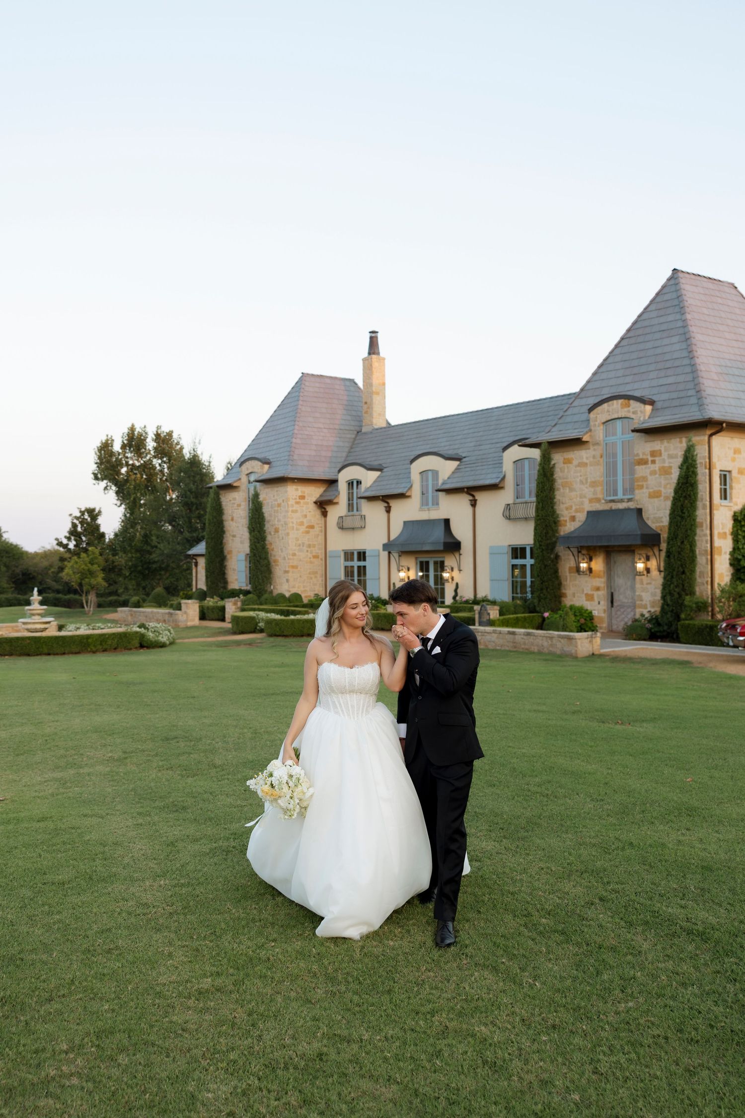 A wedding couple poses on a manicured lawn in front of an elegant French country-style stone mansion.