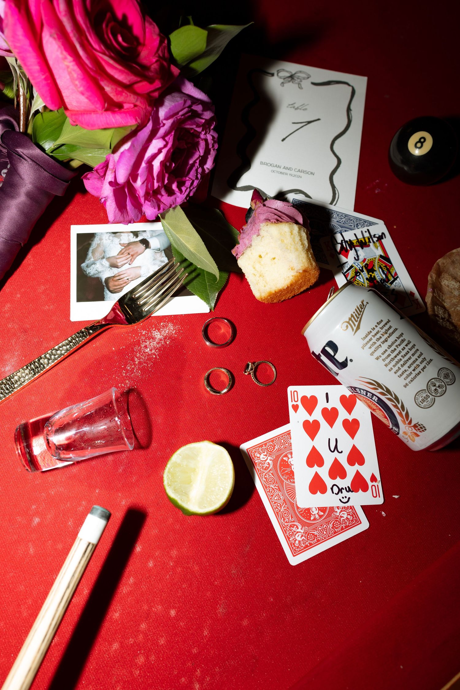 Wedding detail flatlay with rings, playing cards, roses and polaroid photos on red background.