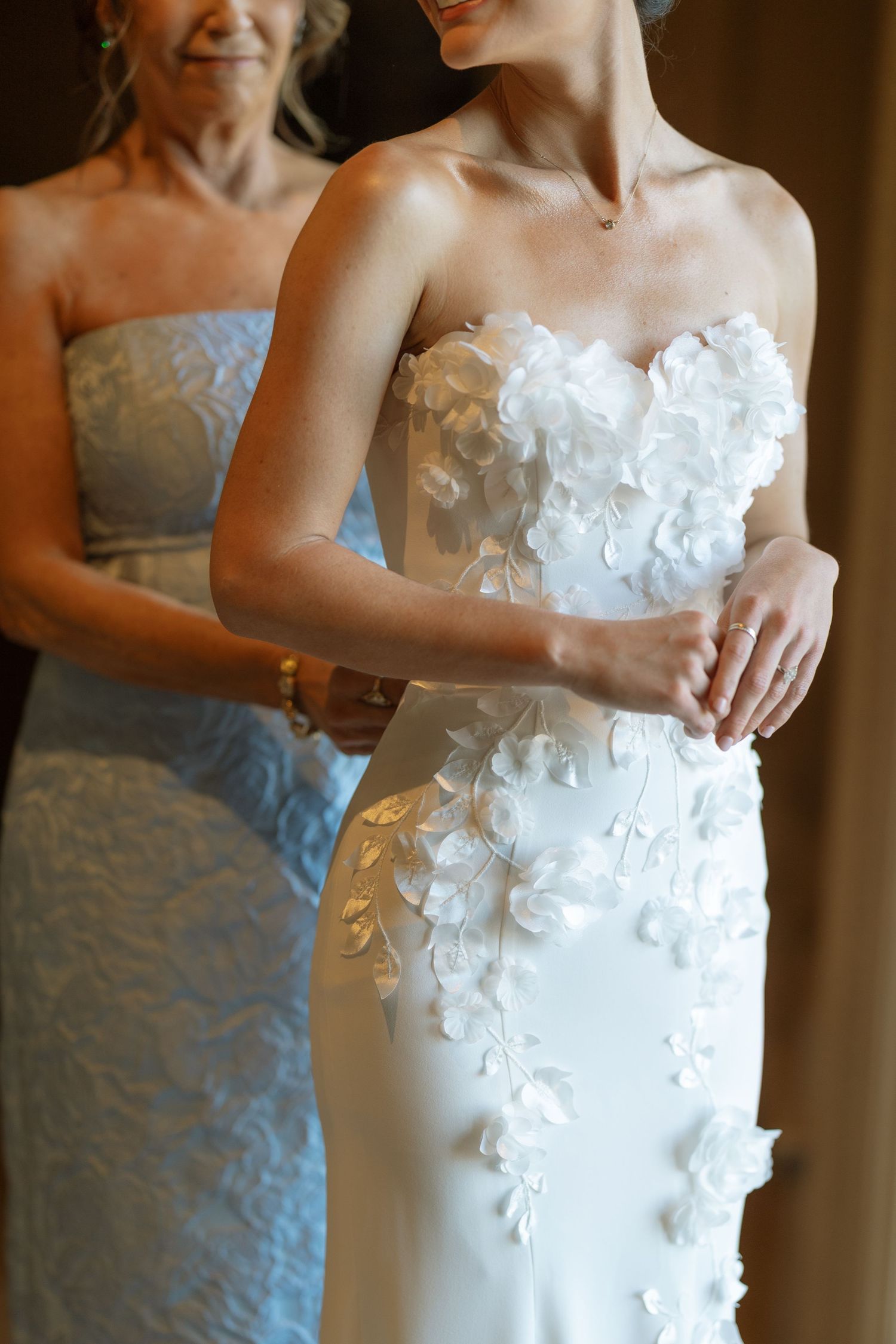 Mother helps bride into strapless white wedding gown with 3D floral applique details.