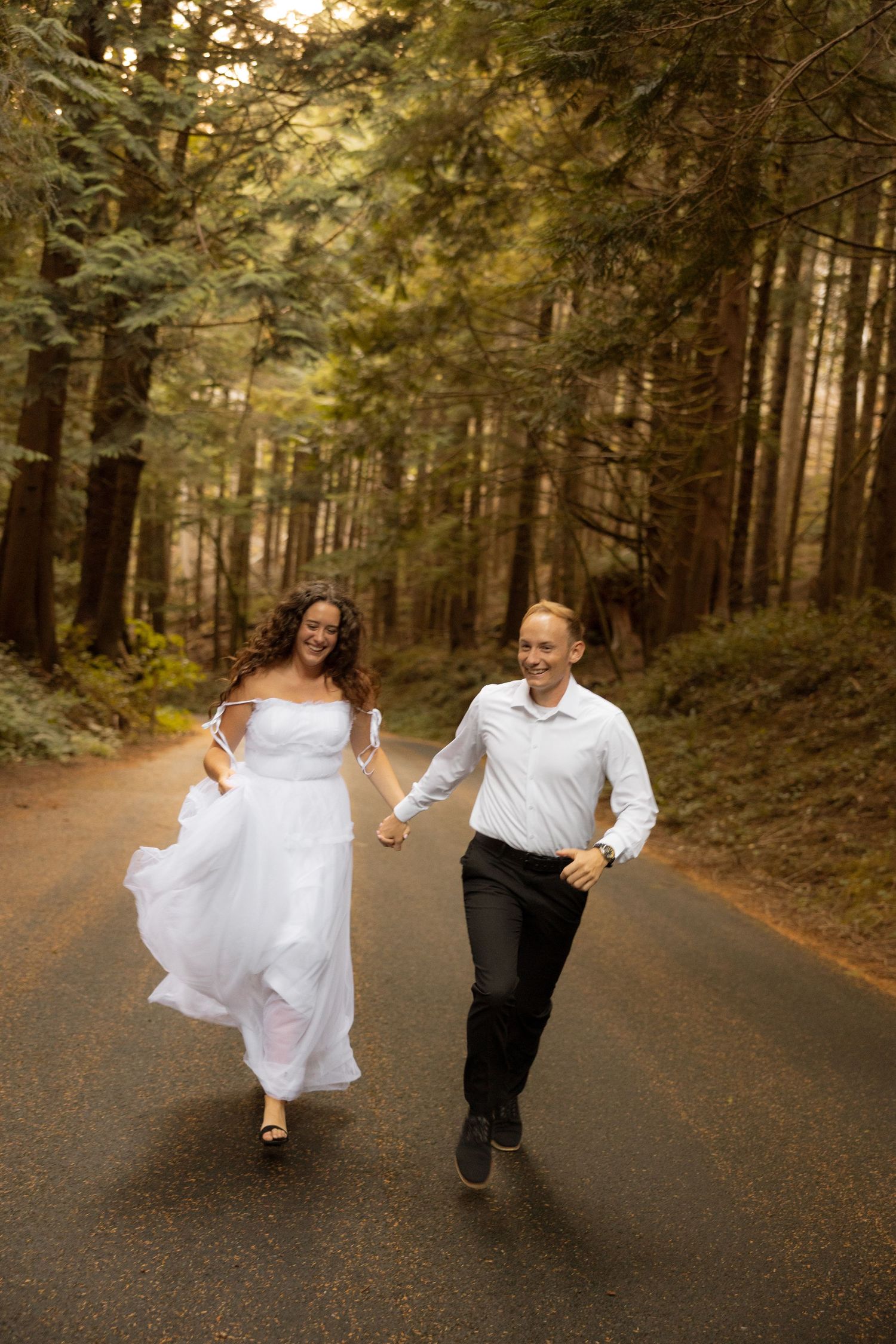 A couple runs joyfully down a forest path surrounded by tall evergreen trees at sunset.