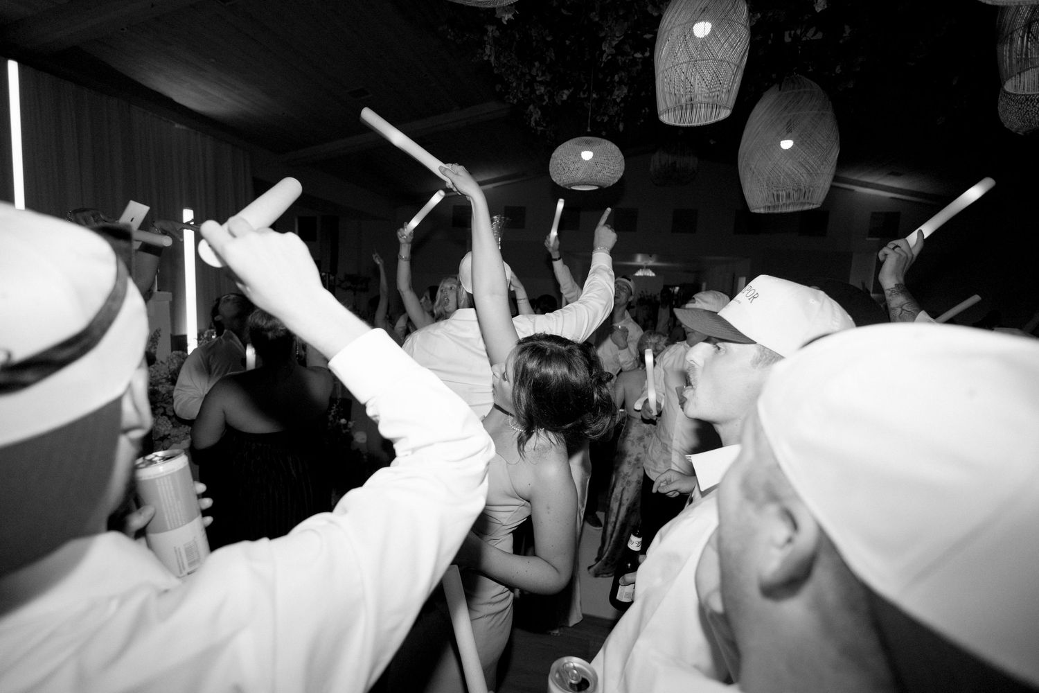 Black and white photo of wedding guests dancing with raised arms under pendant lights at reception.