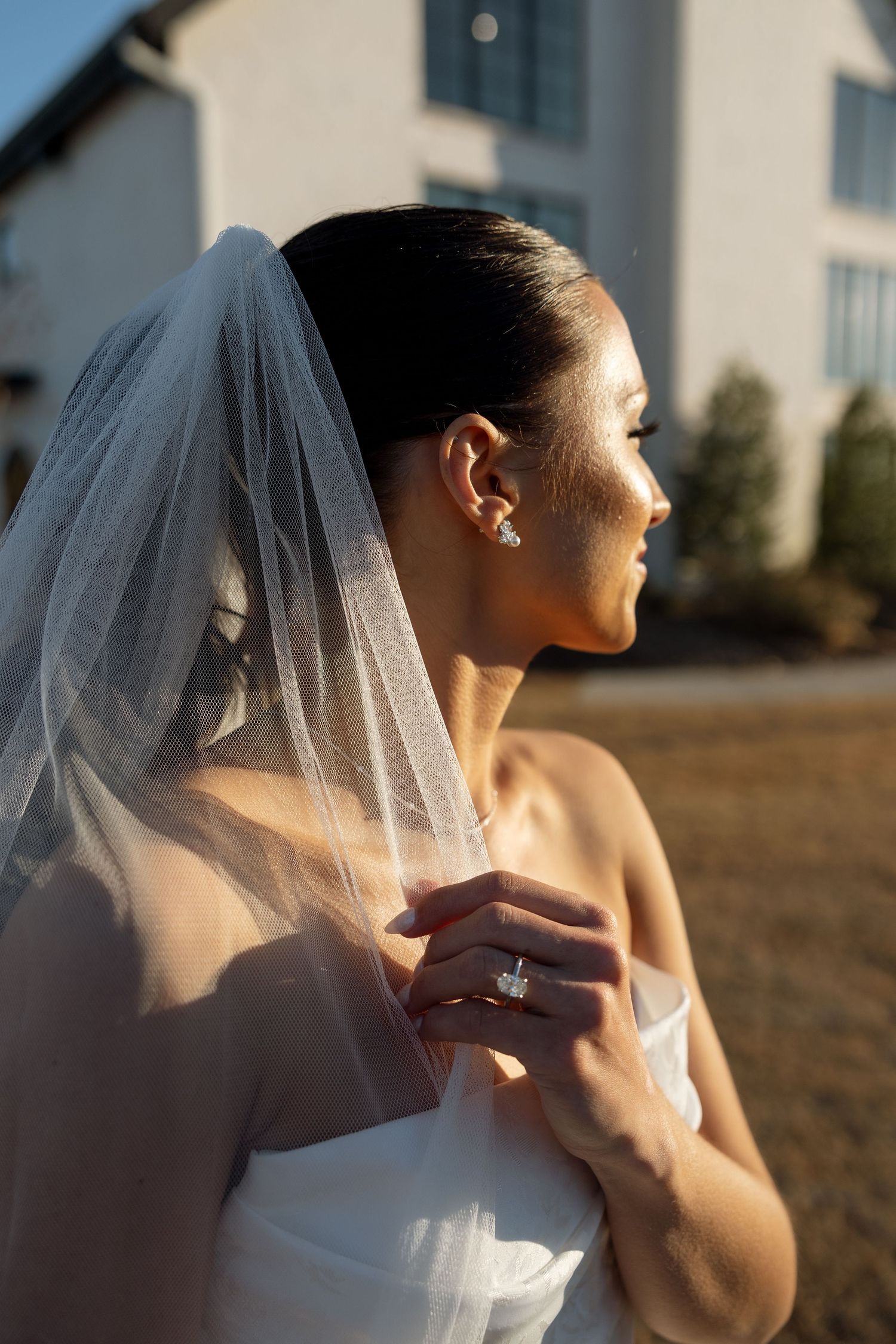 Side profile of a bride in a strapless gown and veil against golden sunlight, showing her sparkling earrings.