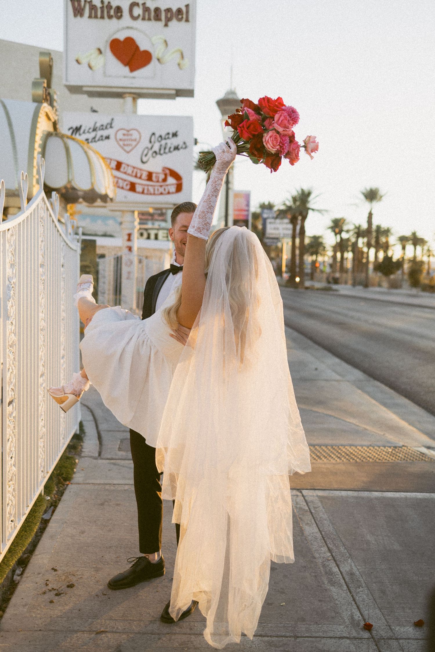 A couple in wedding attire pose outside the Little White Chapel in Las Vegas with palm trees in the background.