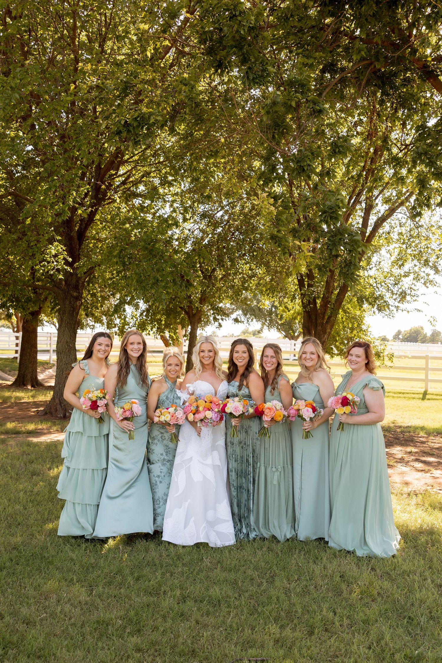 Bridal party in sage green dresses poses together under trees in outdoor setting.