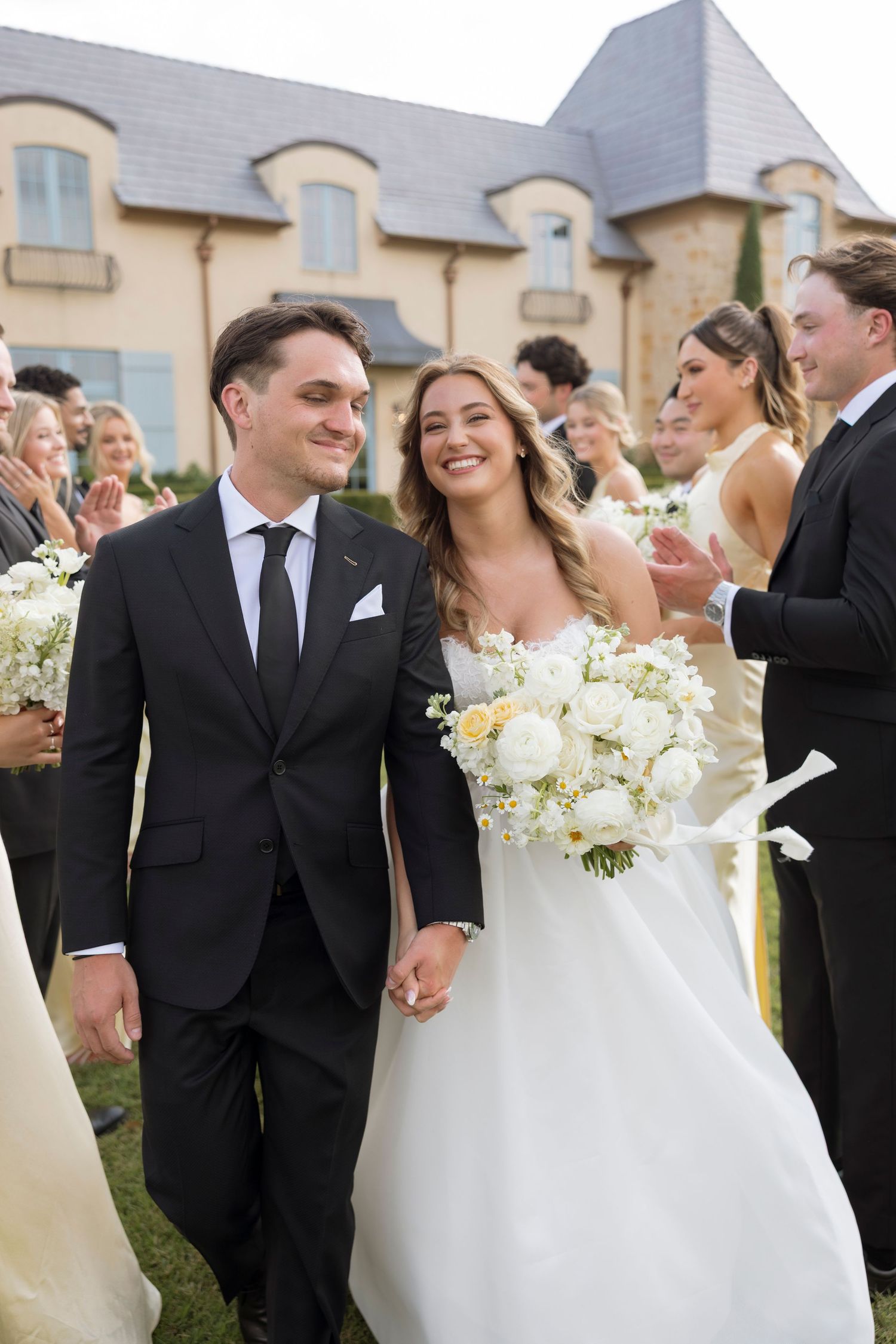 A joyful wedding celebration outdoors with a couple walking together in formal attire against a French-style mansion backdrop.