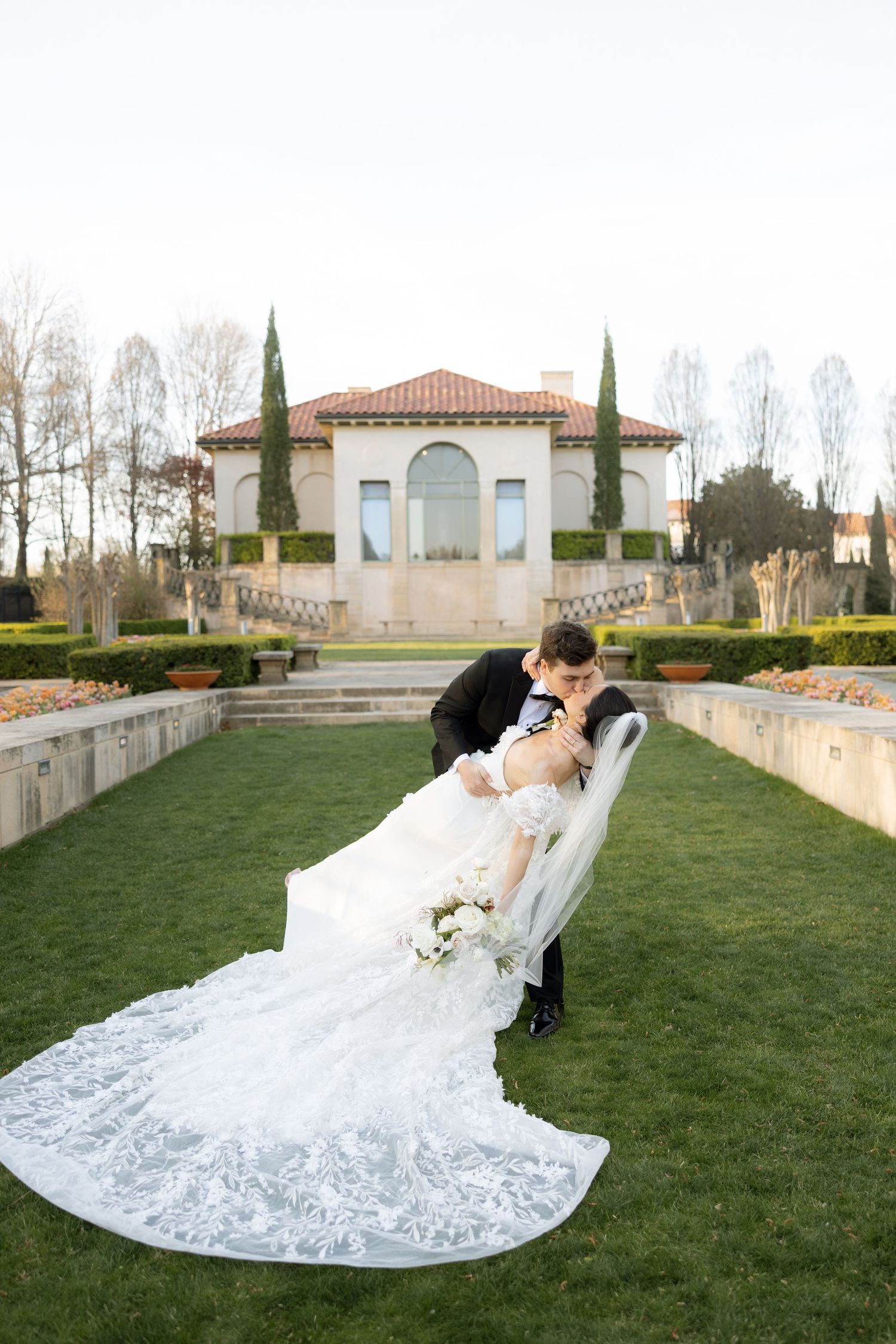 Groom dips bride in lace gown with long train on green lawn before Italian villa.
