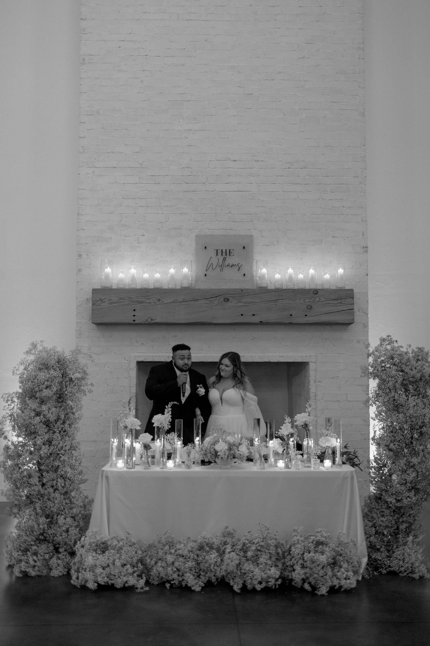 A couple stands behind their beautifully decorated sweetheart table adorned with candles and white flowers.