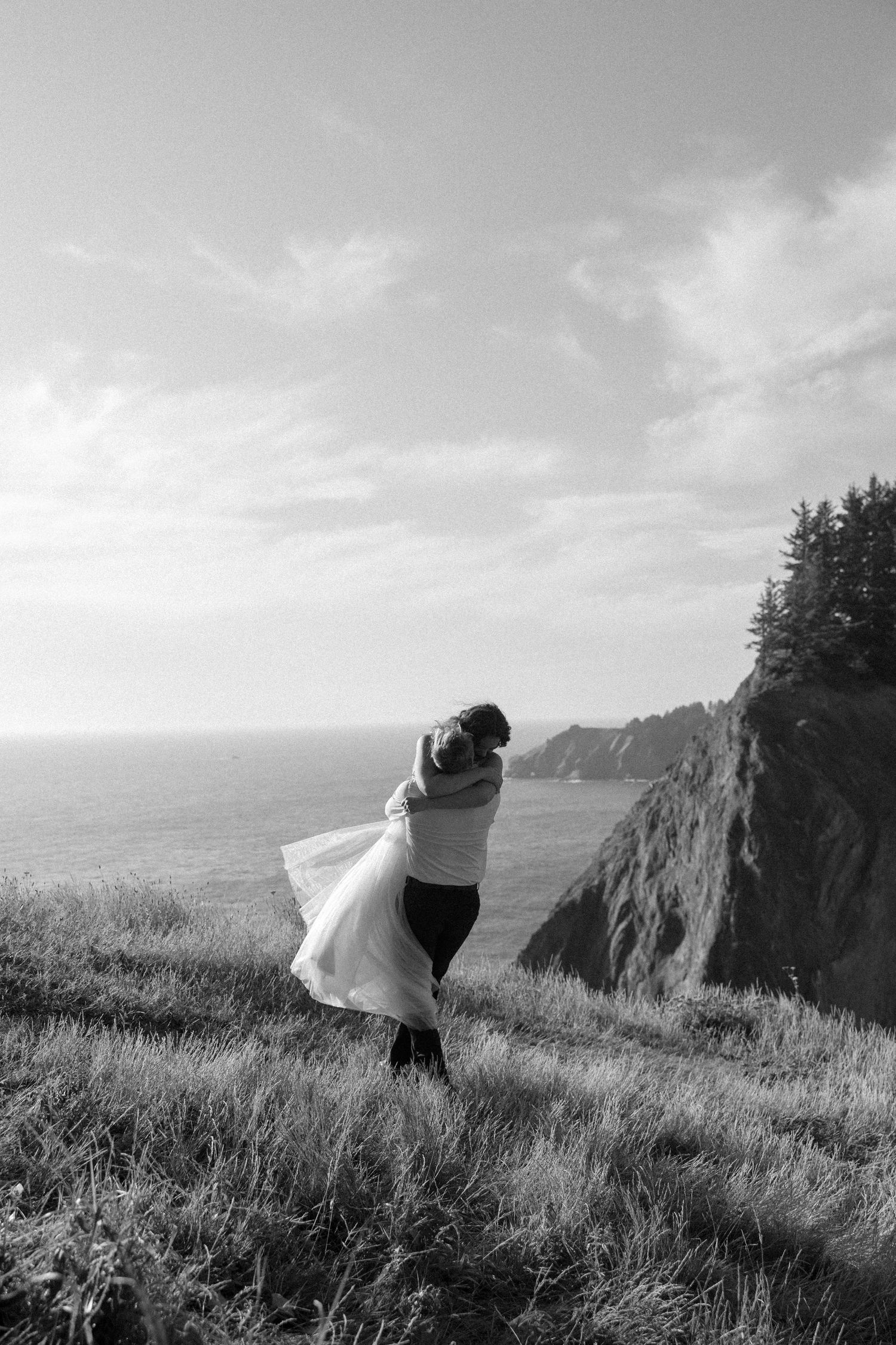 A black and white photo captures a couple dancing on a windswept coastal cliff.