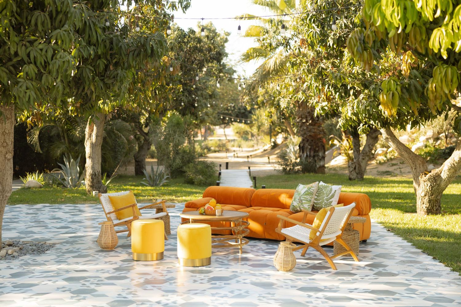 Vibrant outdoor lounge area with orange sofa, yellow chairs, and colorful cushions set among lush garden trees.