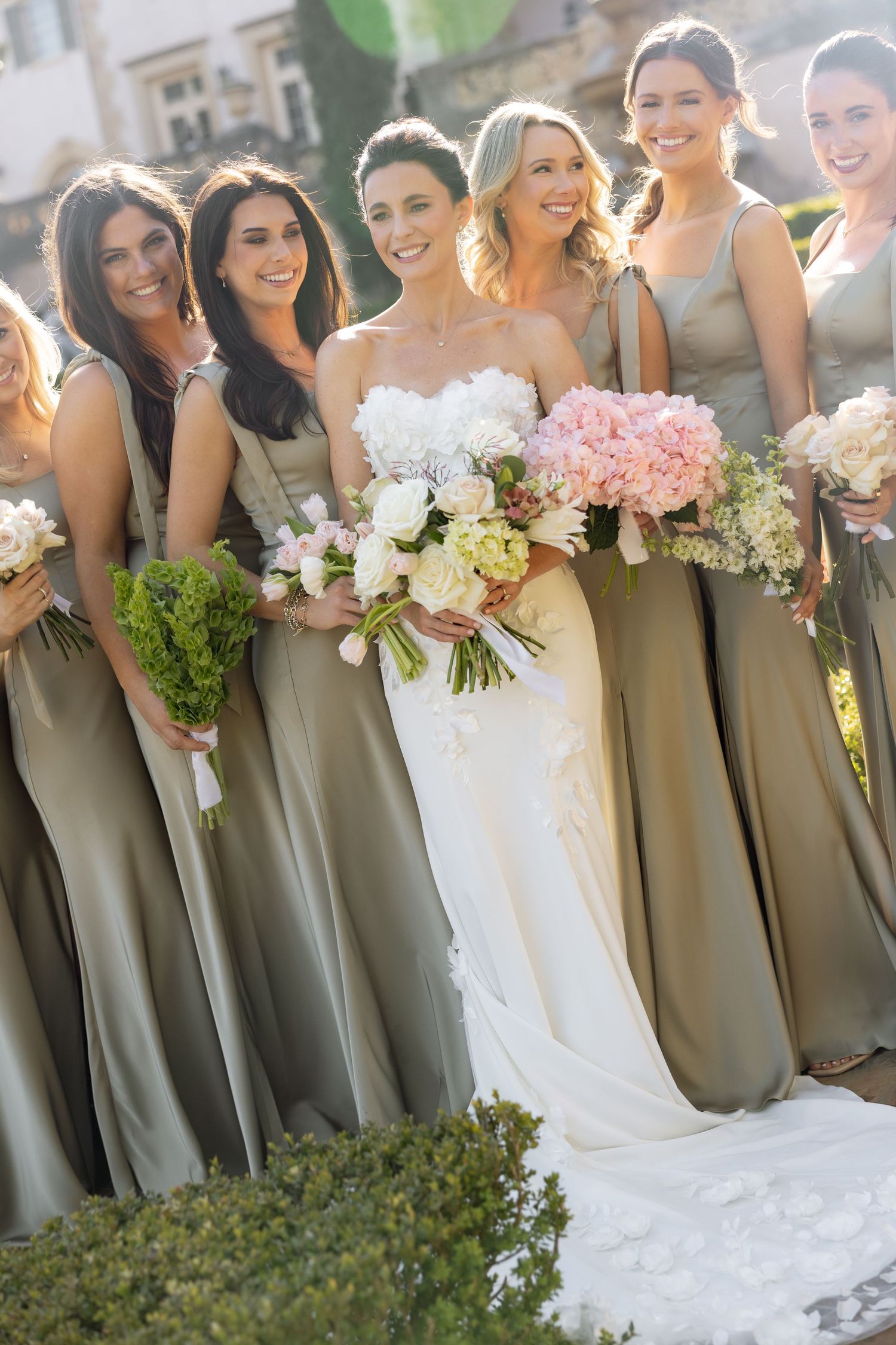 Bride in white strapless gown surrounded by smiling bridesmaids in olive satin dresses holding bouquets.
