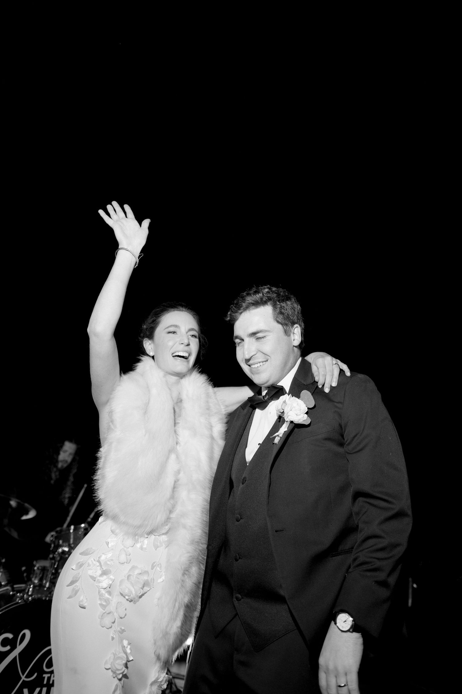 Joyful bride in fur stole raises hand as groom in tuxedo smiles at outdoor wedding reception, black and white photo.