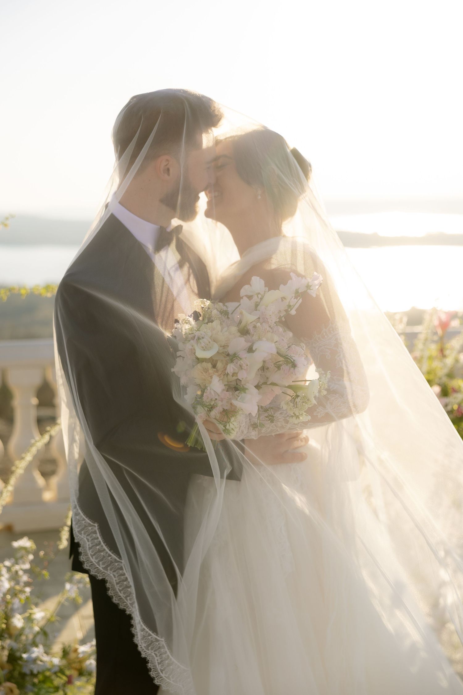 Bride and groom sharing romantic moment under flowing veil, golden backlight, outdoor wedding with white bouquet.