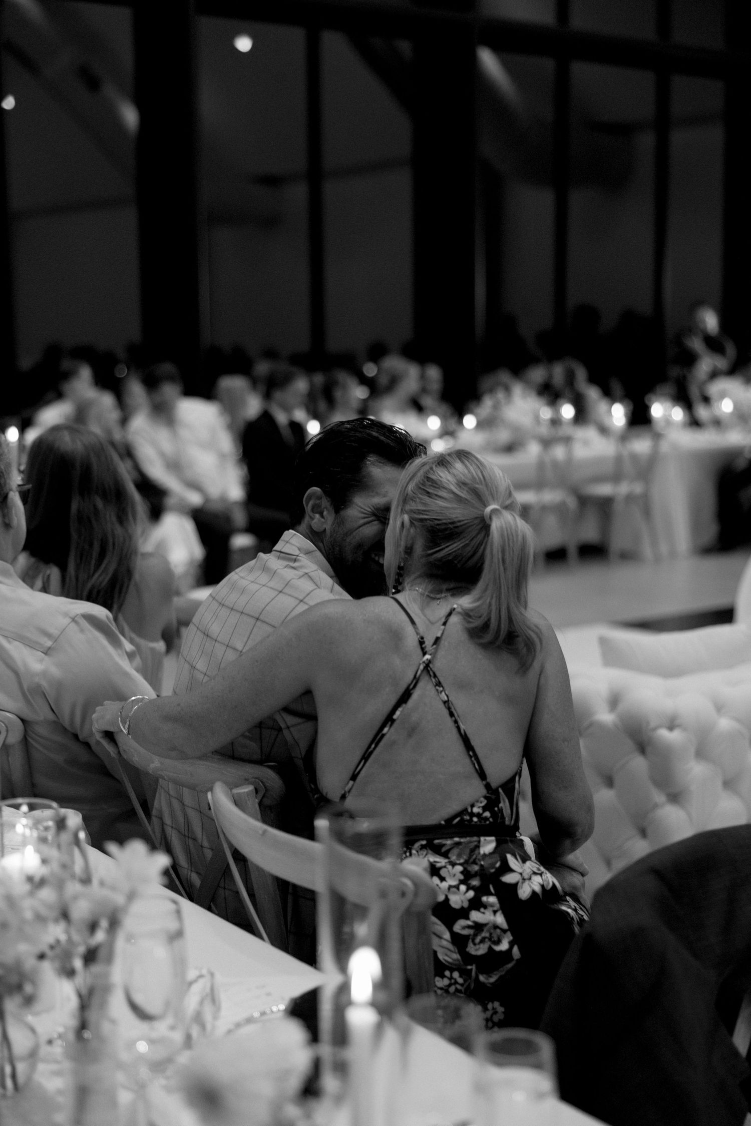 Wedding reception guests seated at elegant tables with white linens and floral centerpieces in a dimly lit venue.