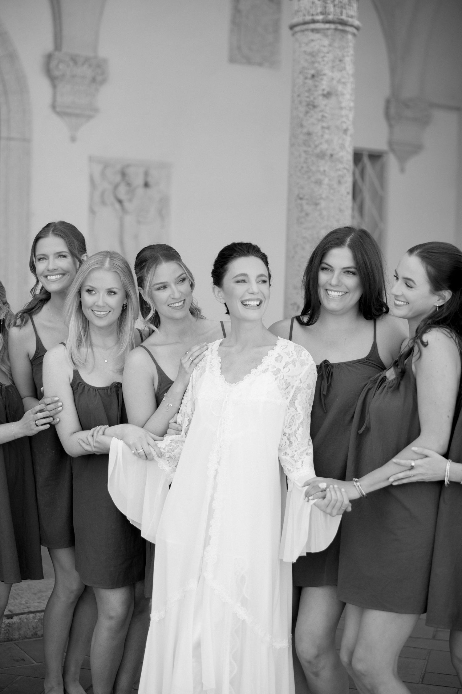 Black and white photo of smiling bride in lace robe surrounded by bridesmaids in short dresses.