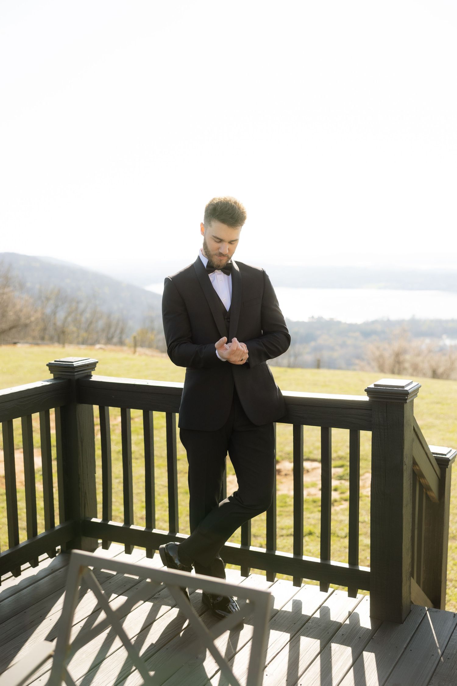 Elegant groom in black tuxedo and bow tie leaning on wooden deck railing with scenic countryside view.