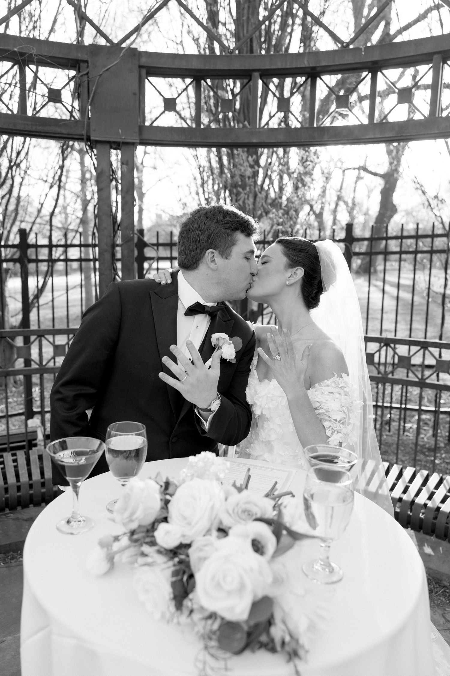 Black and white photo of bride and groom kissing at sweetheart table in romantic iron gazebo.