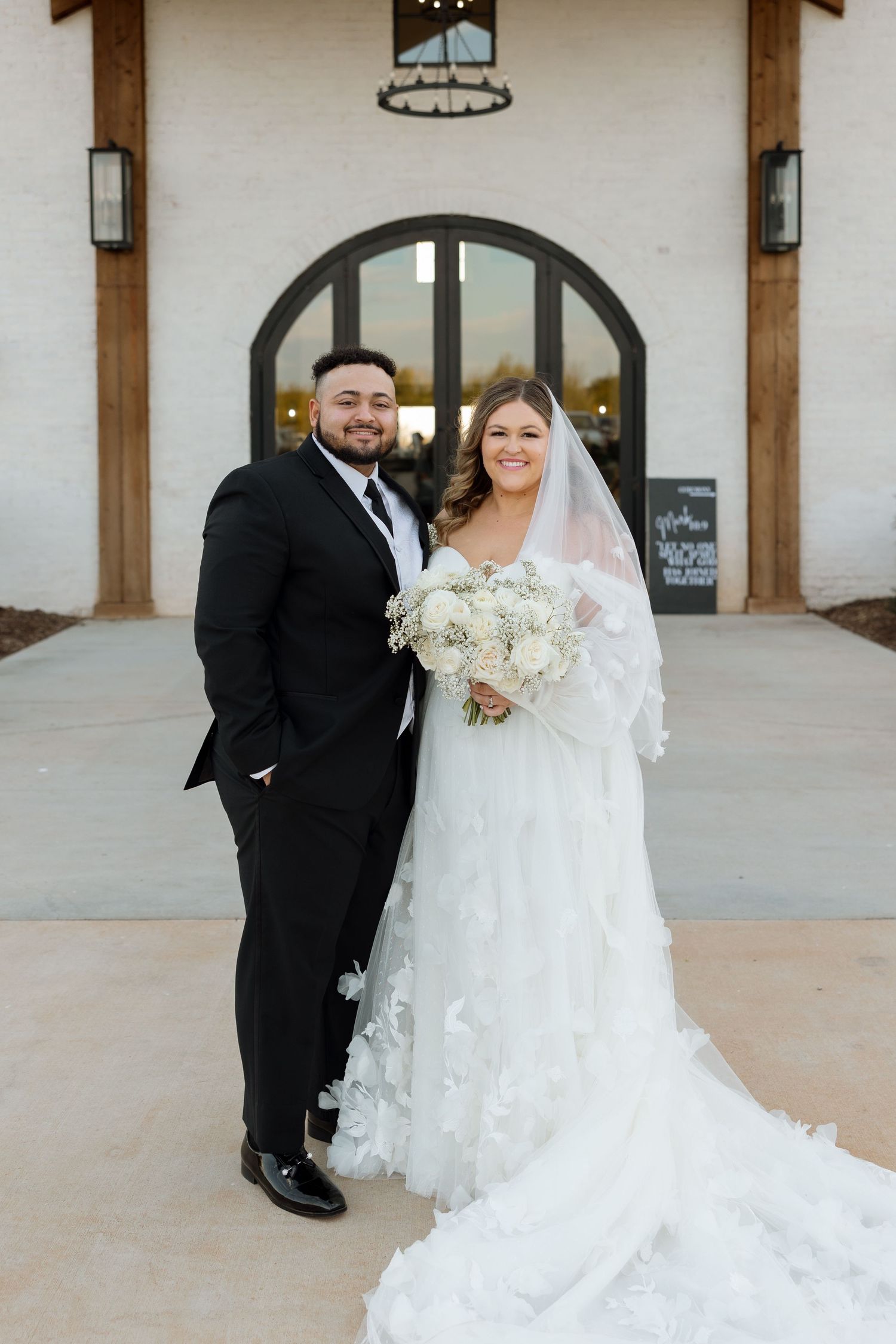 A traditional wedding portrait in front of a church entrance with white flowers and elegant attire.